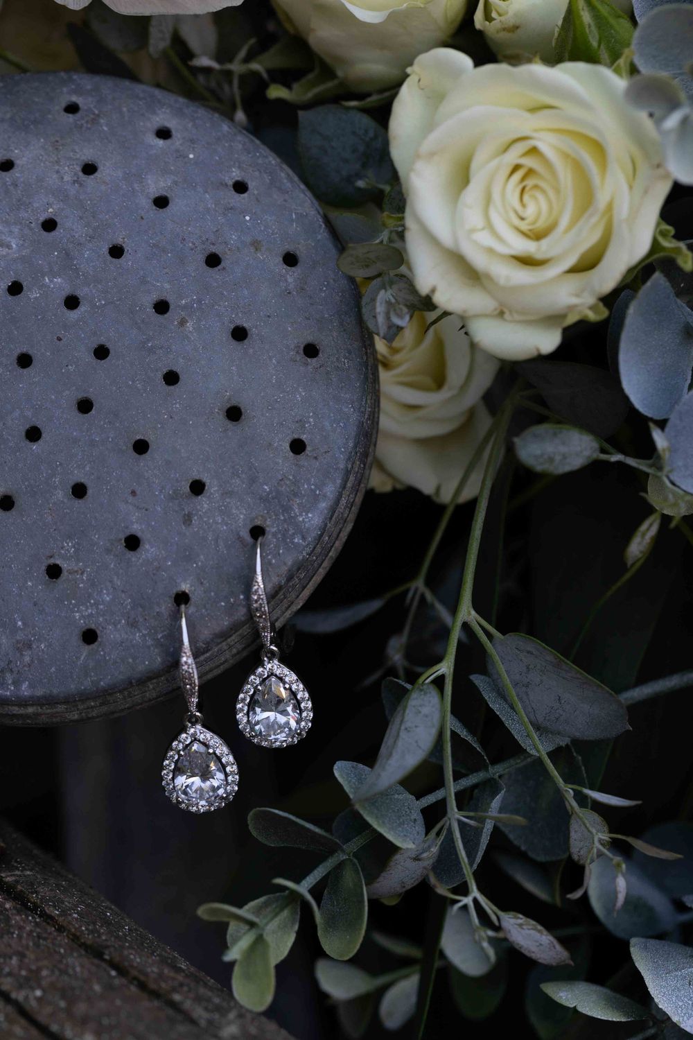 Teardrop crystal earrings displayed with white roses and eucalyptus against a dark perforated surface.