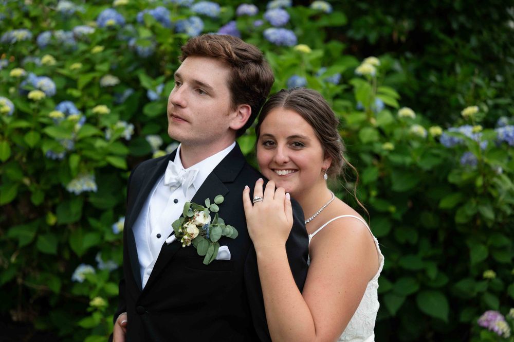 A couple in formal wedding attire posing outdoors with blue hydrangeas in the background.