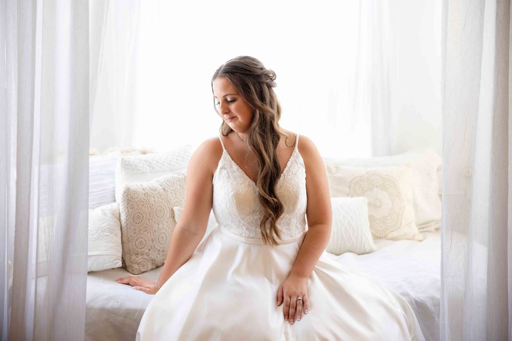 A bride in a white satin wedding gown sits gracefully by a sunlit window surrounded by cream pillows.