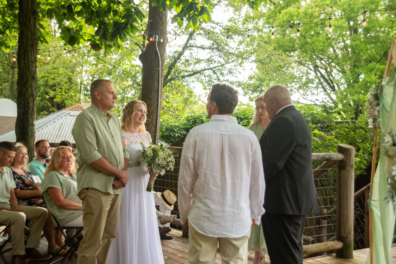 An outdoor wedding ceremony takes place under a green leafy canopy in a garden setting during a sunny day.