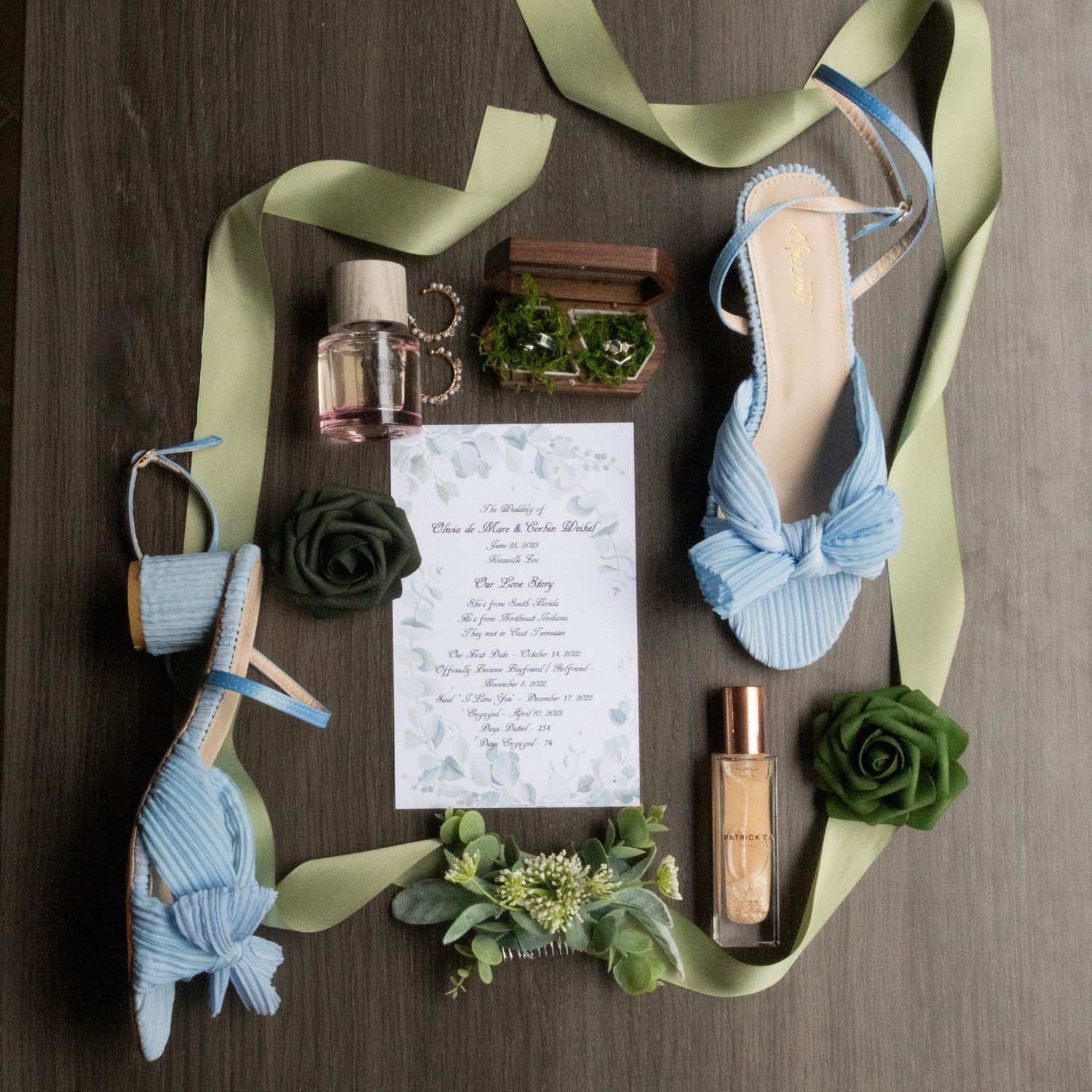 Wedding accessories flatlay with light blue ribbon heels, green succulents, perfume bottle and handwritten invitation note.