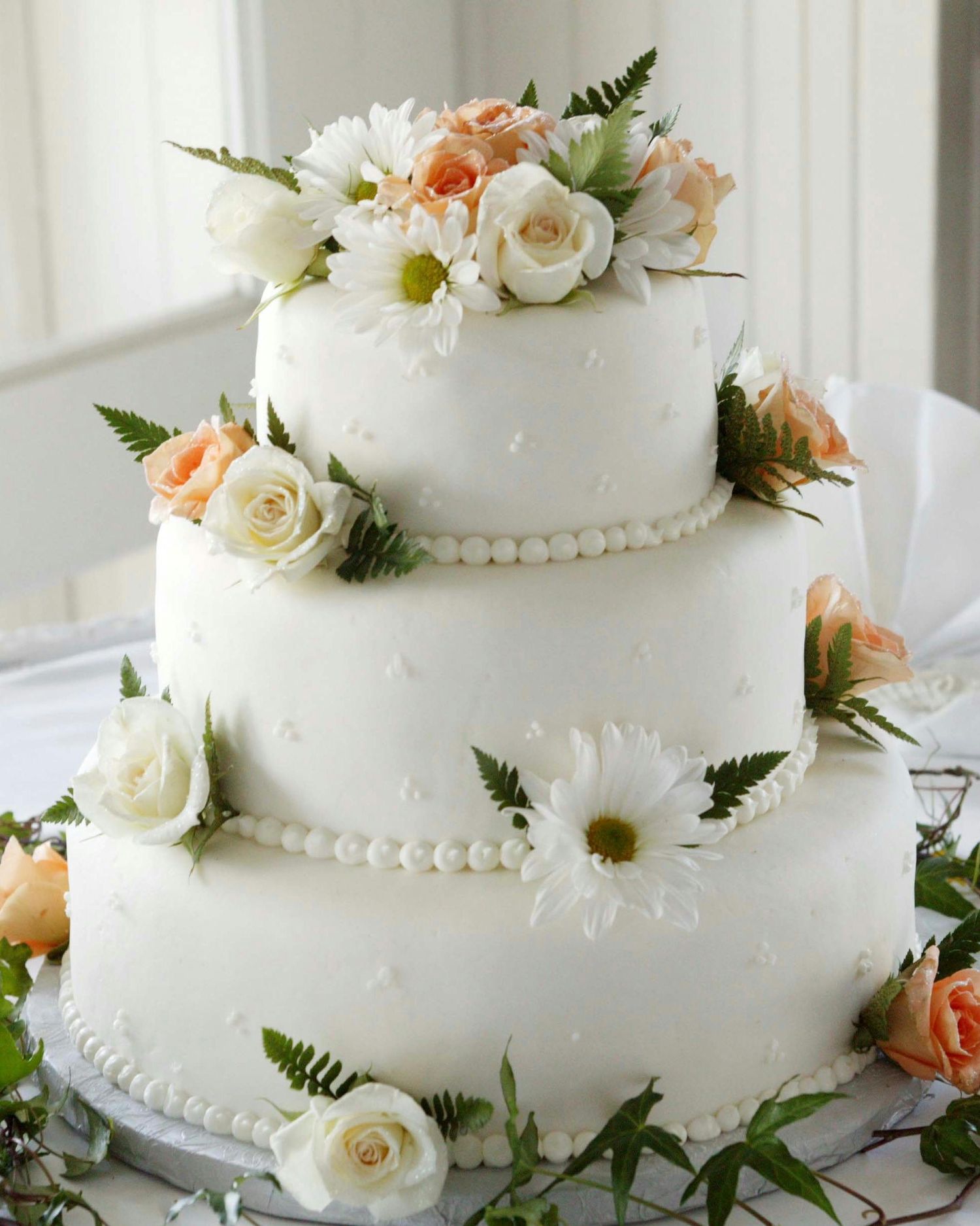 Three-tiered white wedding cake decorated with fresh peach and ivory roses, white daisies, and delicate pearl borders.