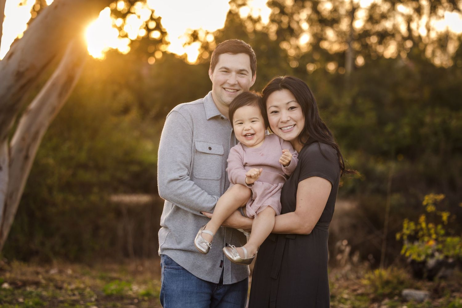 A family stands together outdoors during golden hour sunset with trees in the background.