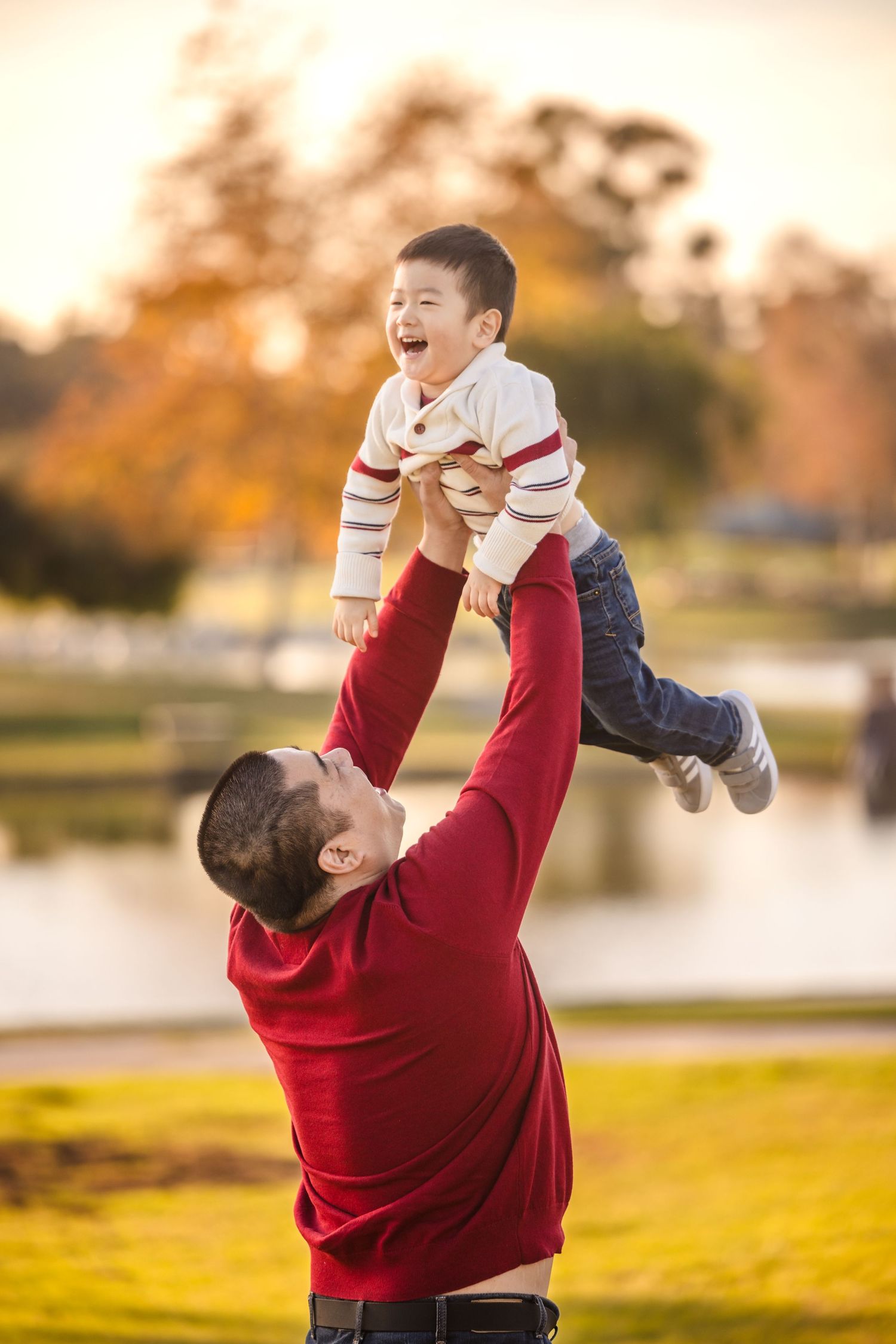 A joyful moment captured at sunset in a park as a person in red lifts a laughing child wearing stripes into the air.