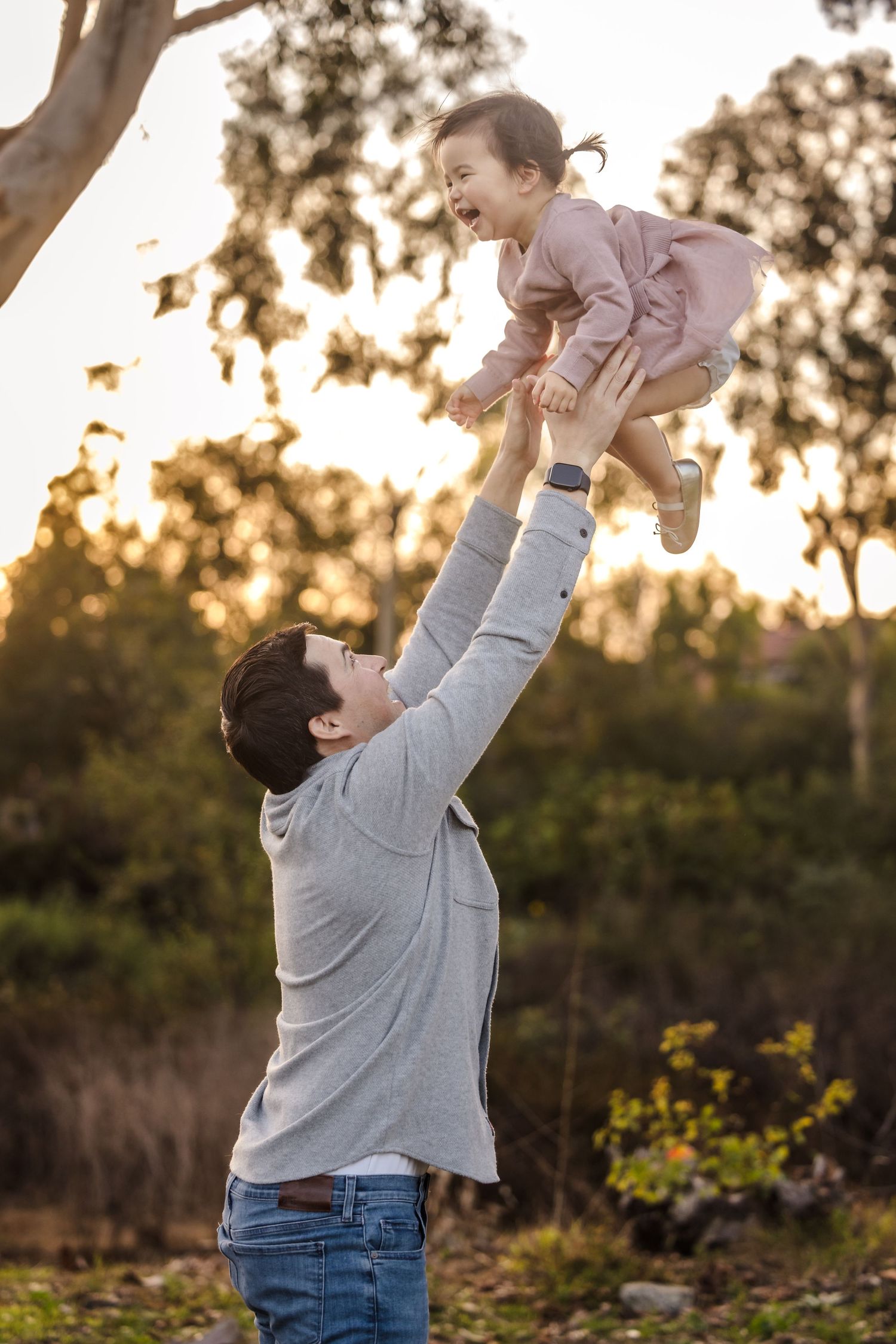 A joyful playtime moment as a child in pink is lifted high into the air at sunset in a park setting.