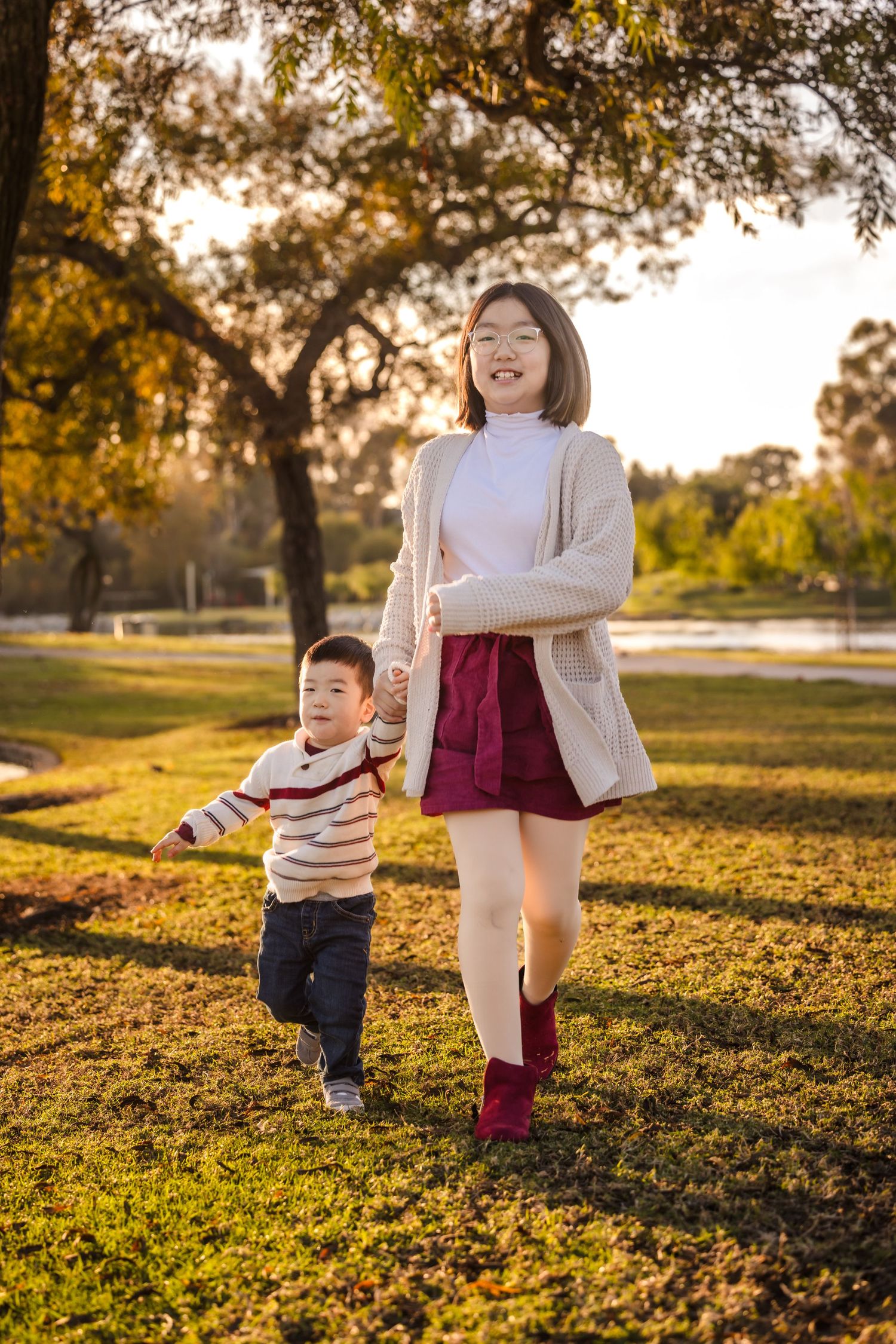 Two people walk together in a park during golden hour sunset with autumn trees and grass in the background.