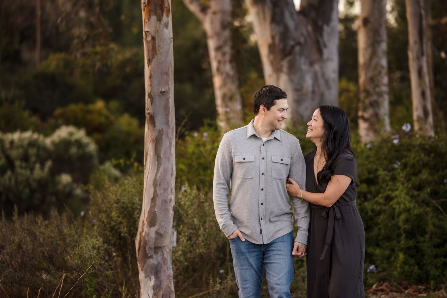 Two people share a tender moment while standing among tall eucalyptus trees in a serene forest setting.