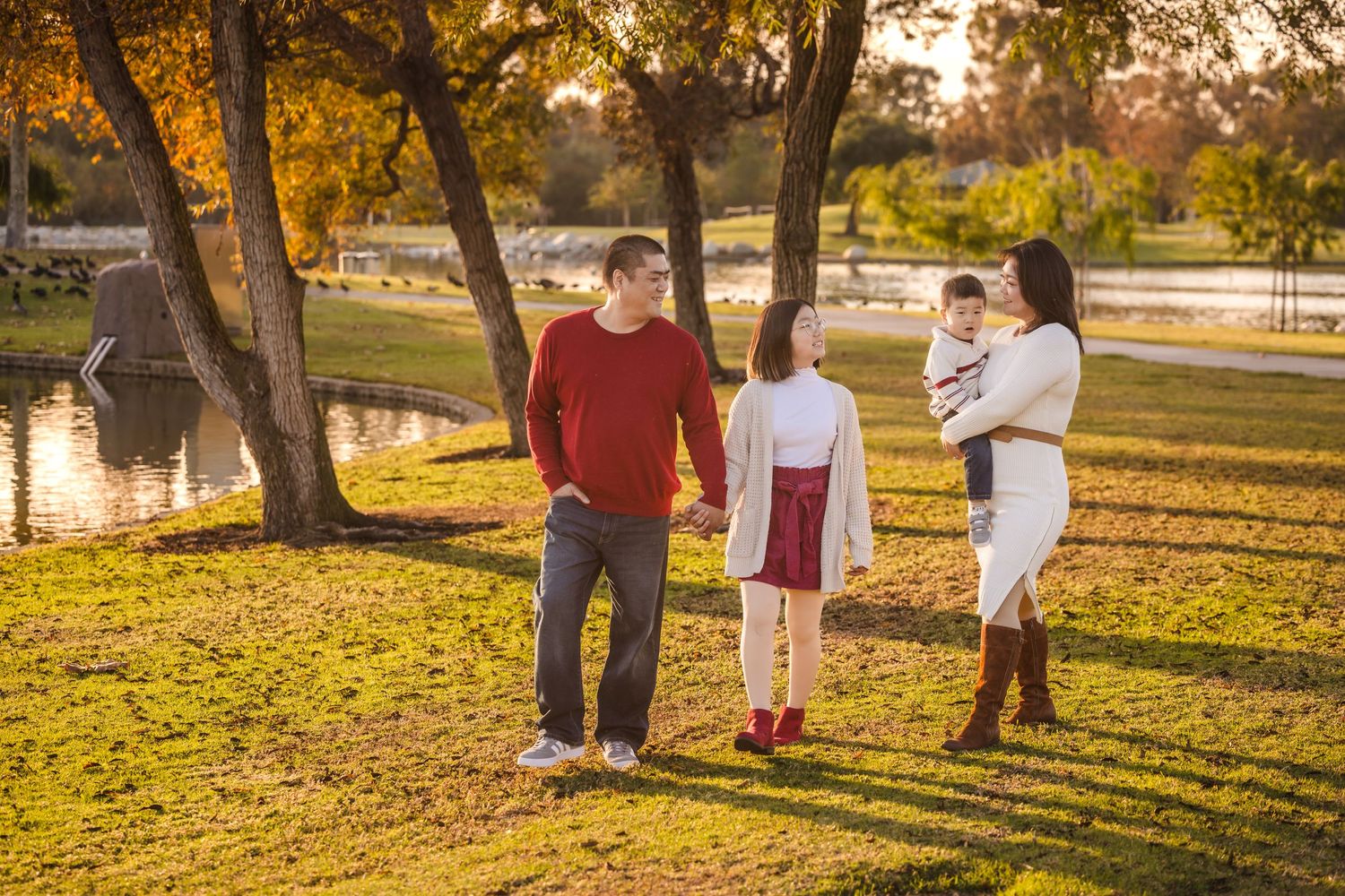 A family enjoying a warm autumn evening in a park with golden sunlight filtering through trees near a pond.