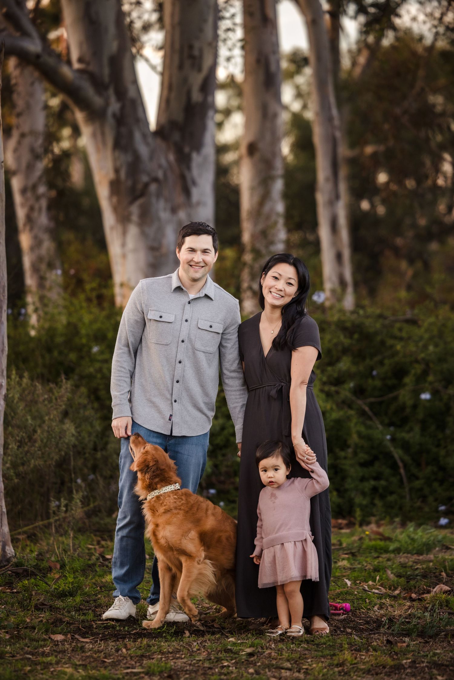 A family stands together in a wooded area with their golden retriever dog at sunset.