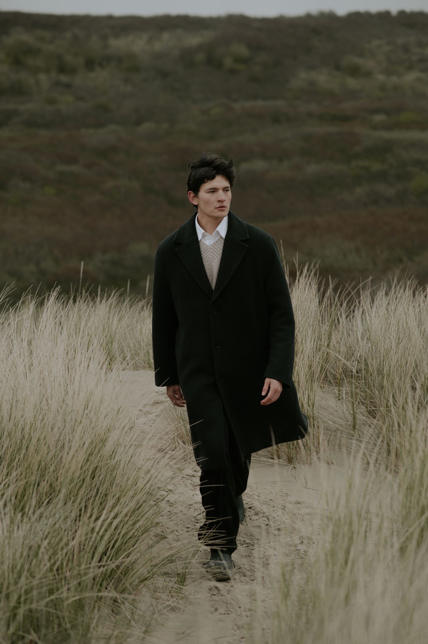 A person in a black coat walks through tall grass in a moody, overcast coastal landscape.