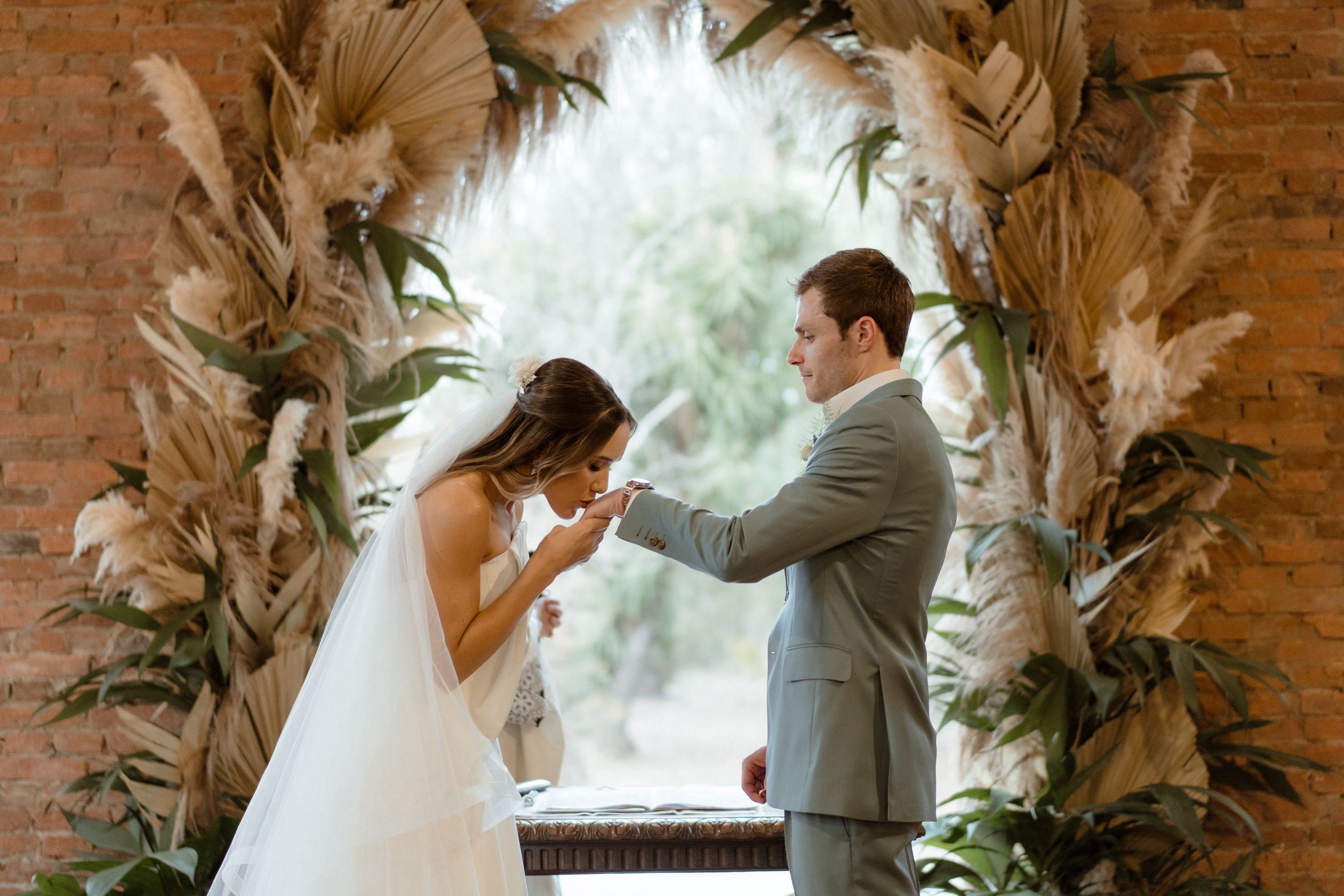 A romantic exchange during a wedding ceremony beneath a dried palm leaf and floral arch.