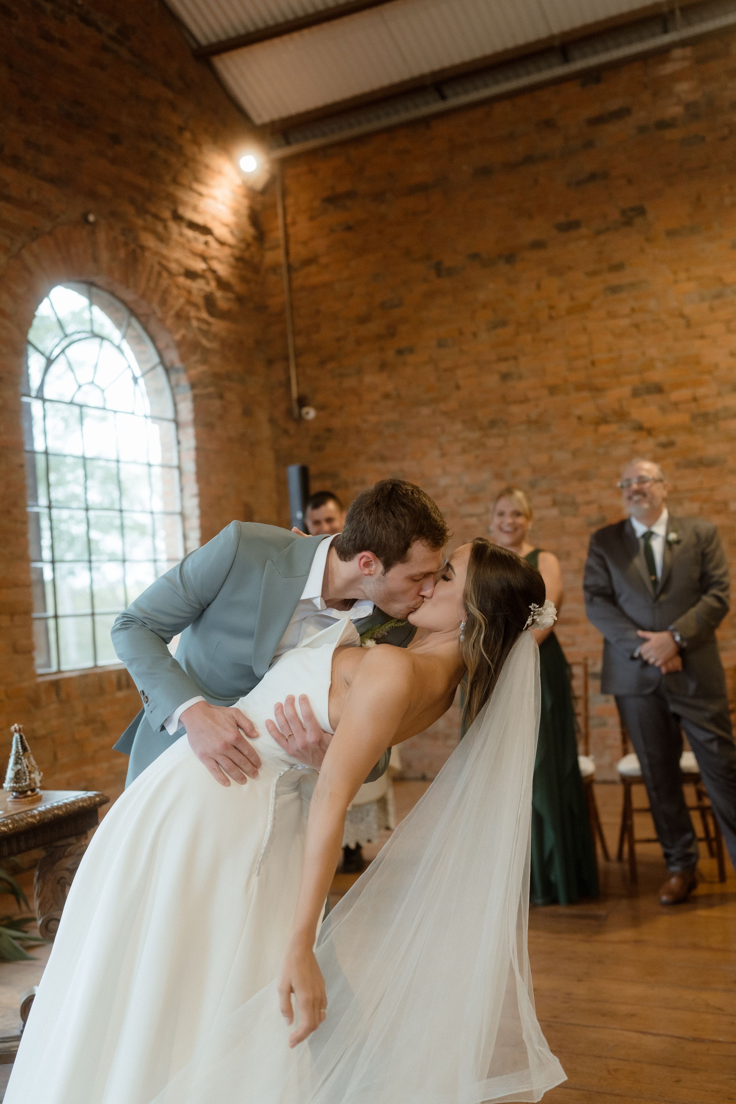 A romantic dip and kiss during a wedding ceremony in a rustic brick venue with arched windows.