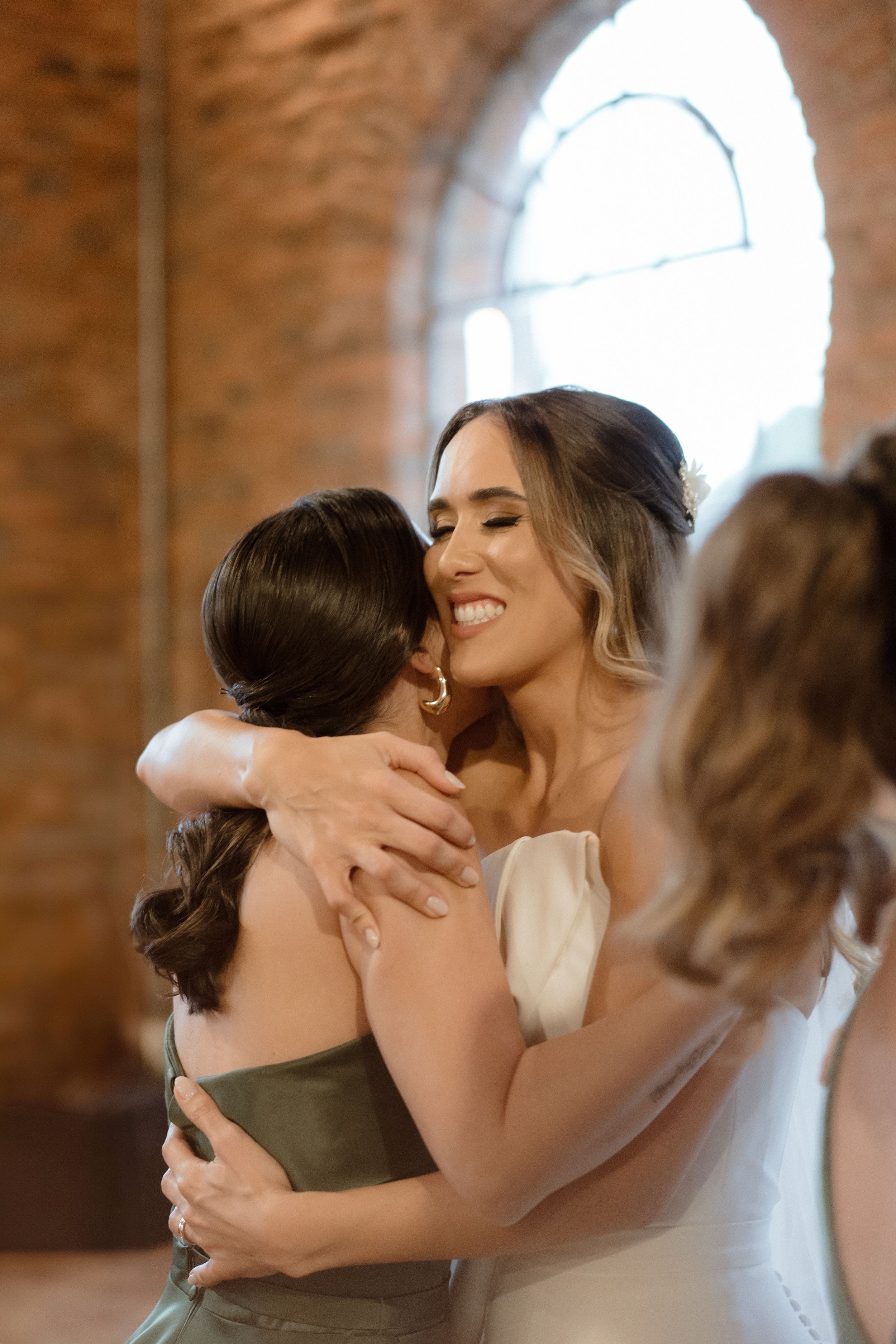 An emotional embrace between the bride and wedding party member in a sunlit brick venue.