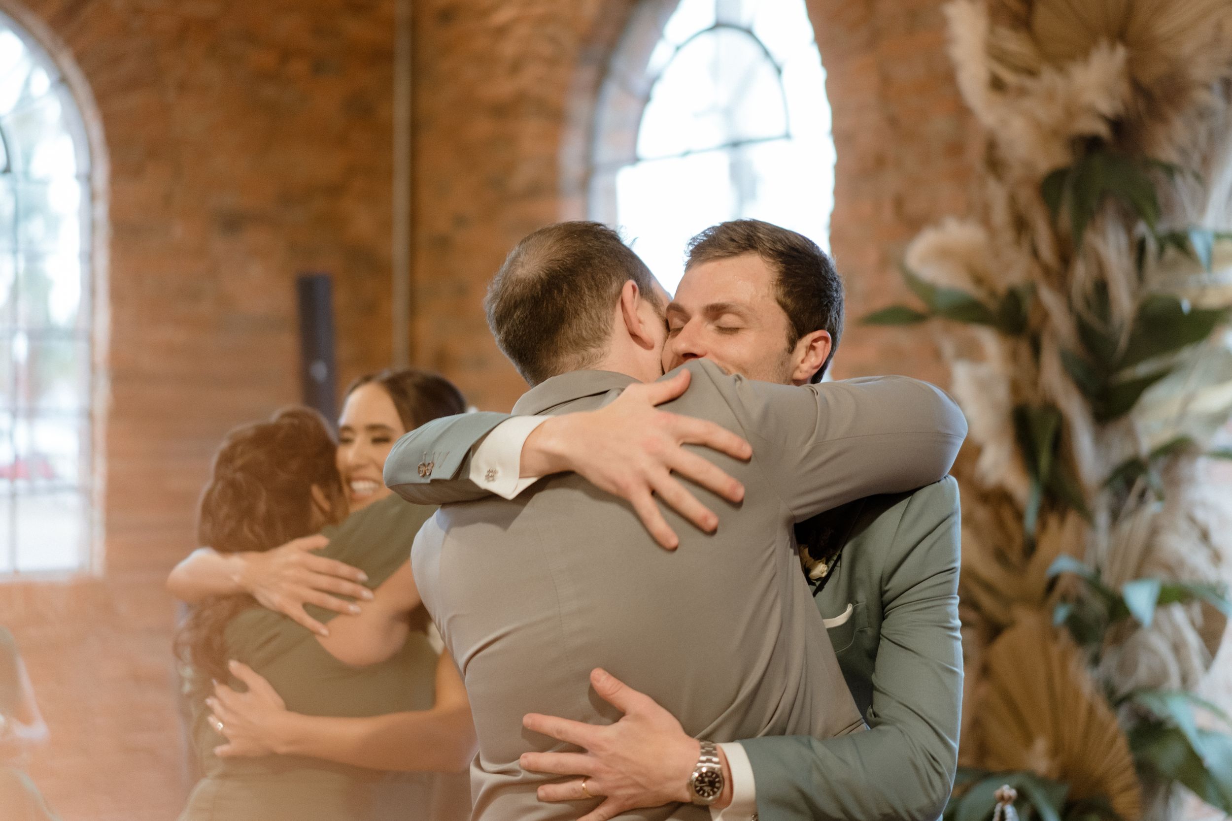 Joyful group hugs being exchanged during wedding celebrations in a rustic brick venue.
