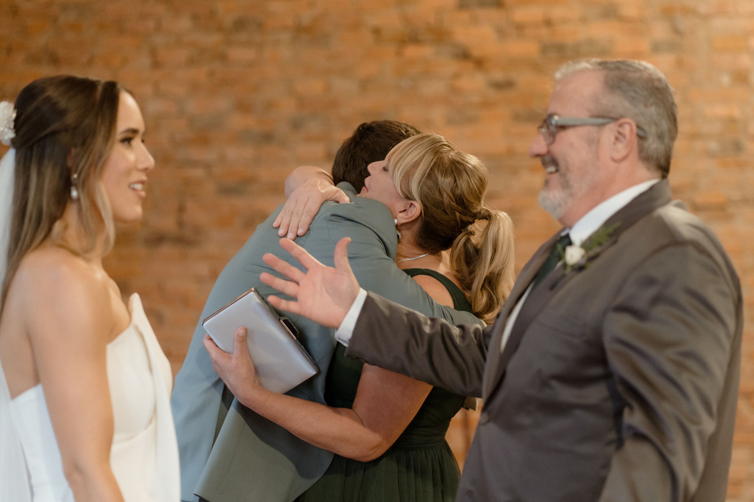 Wedding party members share emotional embraces while others look on in a brick-walled venue.