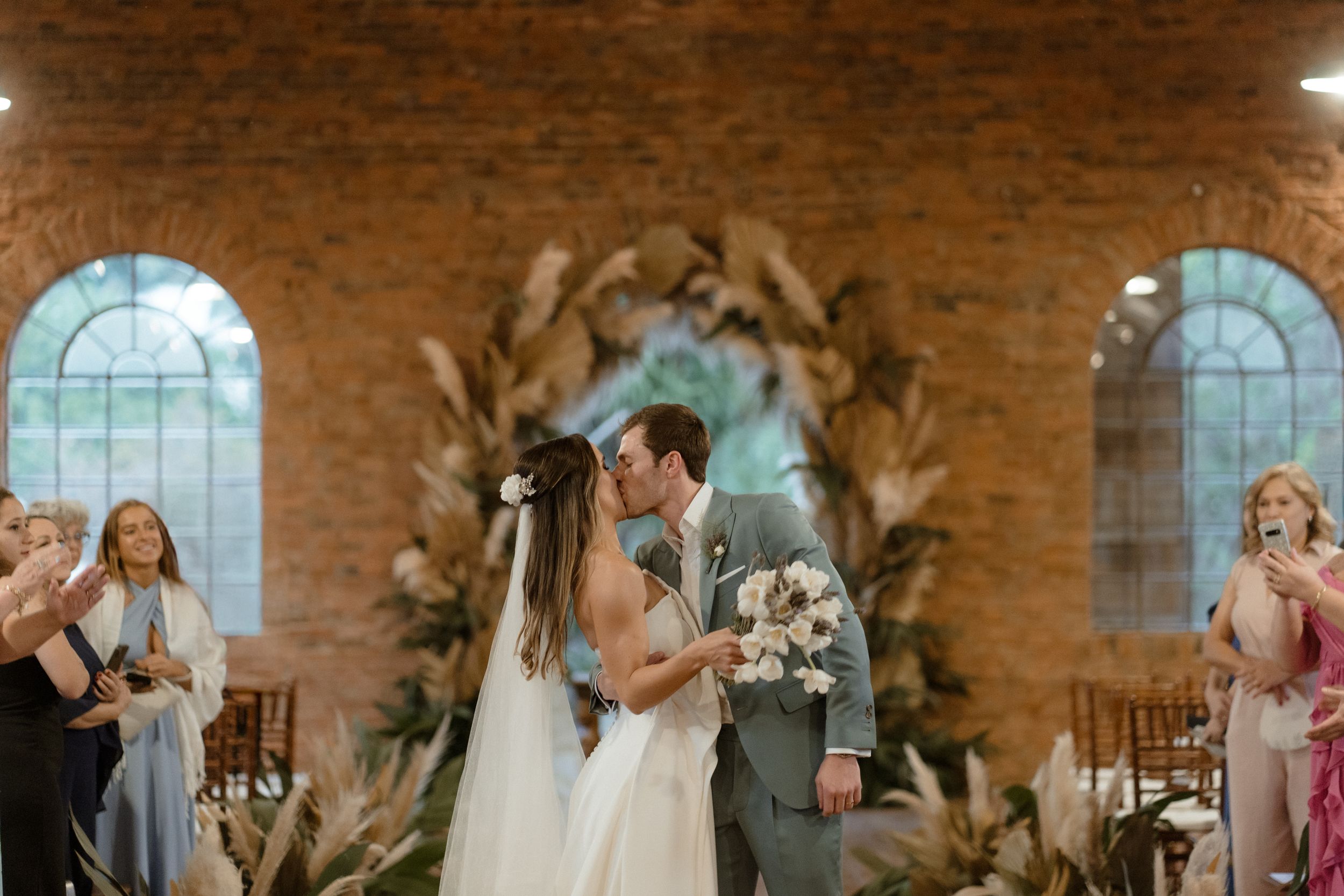 A wedding ceremony in a rustic brick venue with arched windows and pampas grass decor.