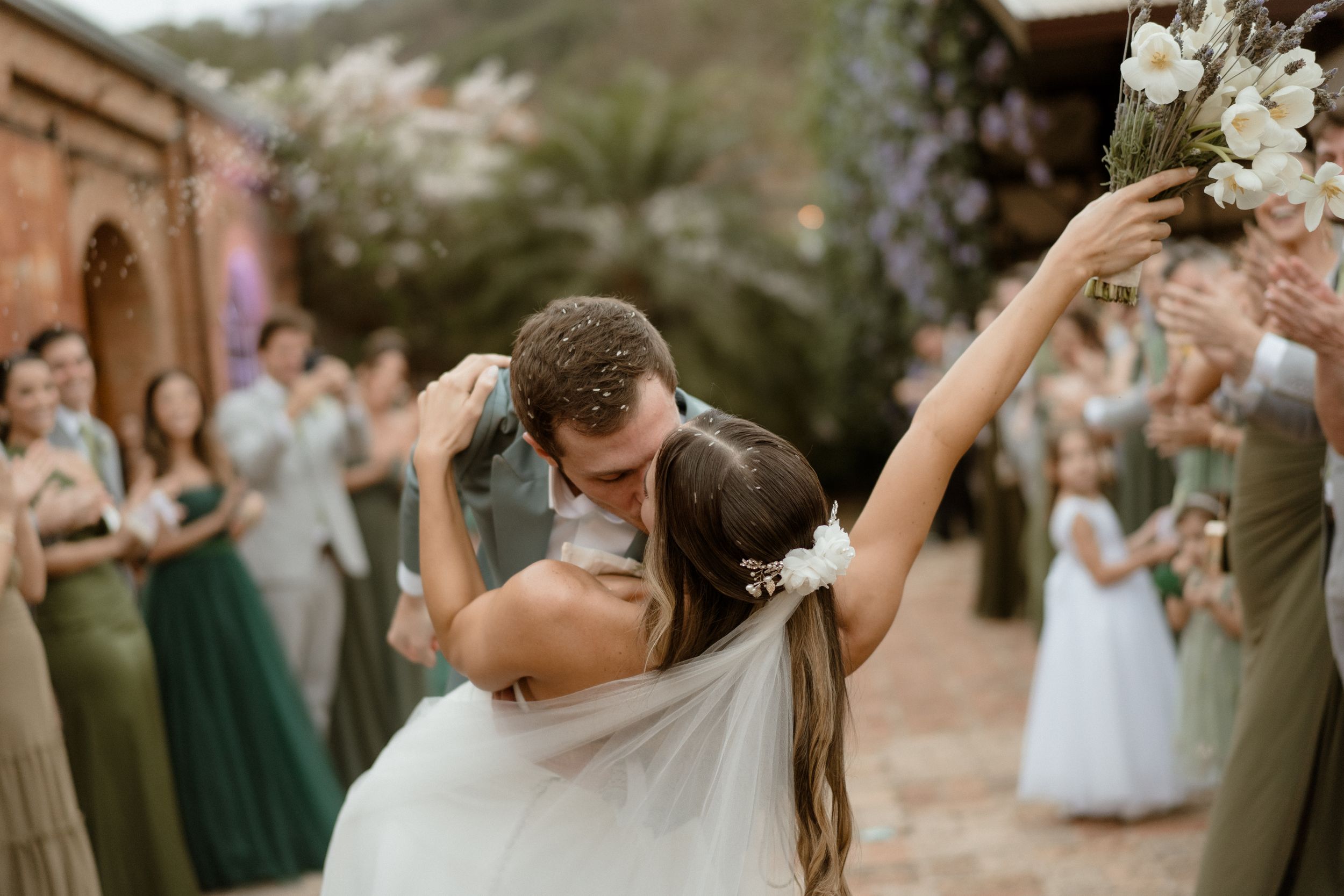 A romantic dip kiss between newlyweds surrounded by bridesmaids in green dresses at an outdoor wedding celebration.