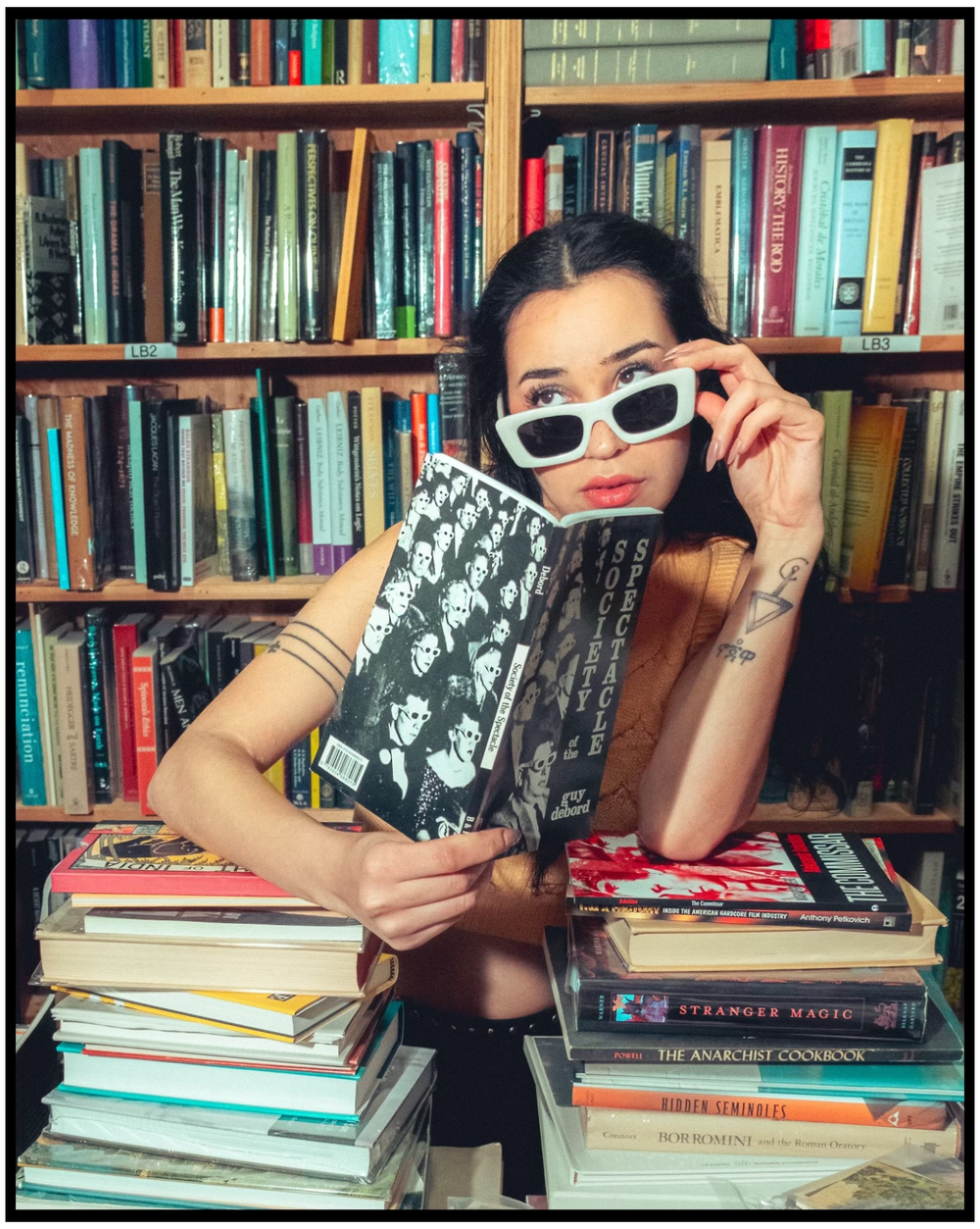 Stylish person in white sunglasses poses with books in front of a colorful library bookshelf. Portland portrait photographer