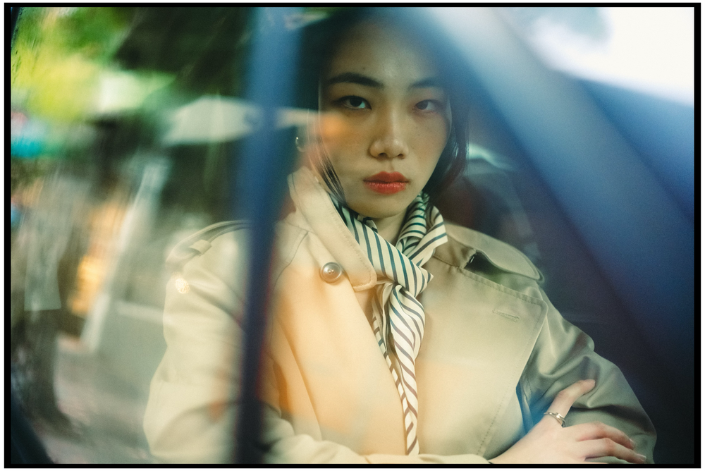 A vintage-style portrait through a car window showing someone wearing a beige coat and striped neck scarf. Portland portrait photographer