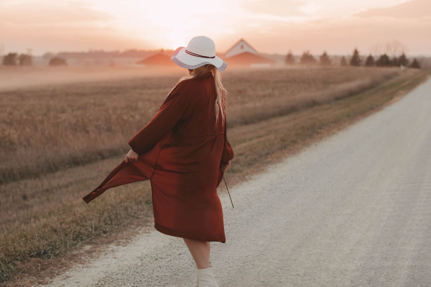Person in a flowing red coat and white hat walks along a dusty rural road at sunset.