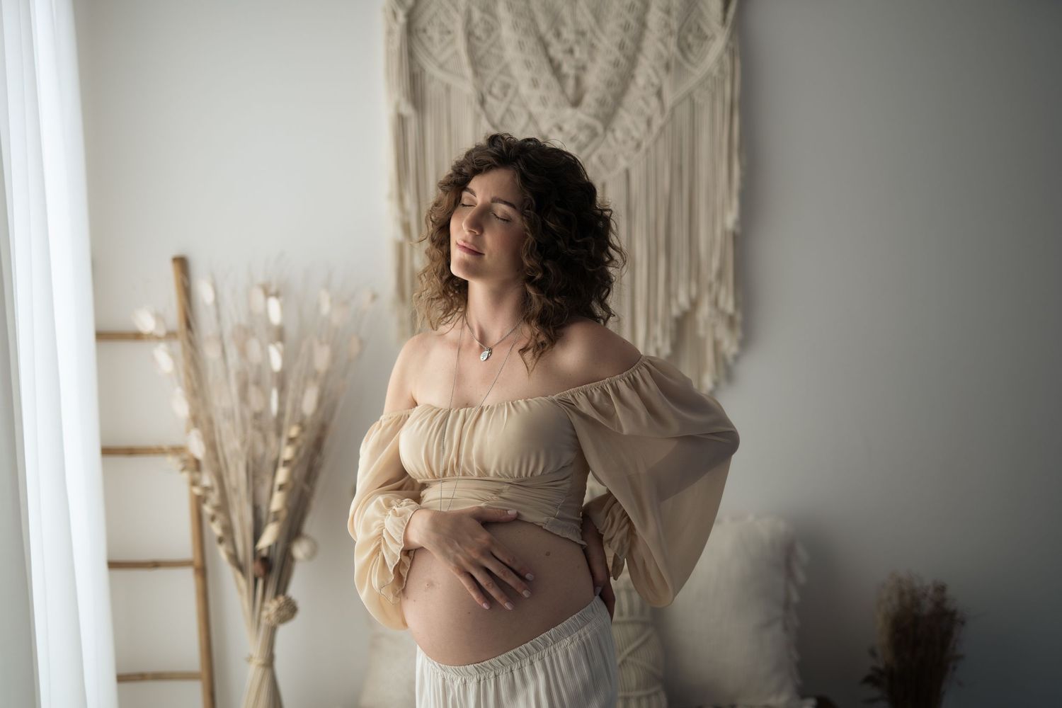 Giovane futura mamma con gli occhi chiusi in completo spezzato beige, accarezza il pancione nudo. Sullo sfondo un set fotografico romantico.