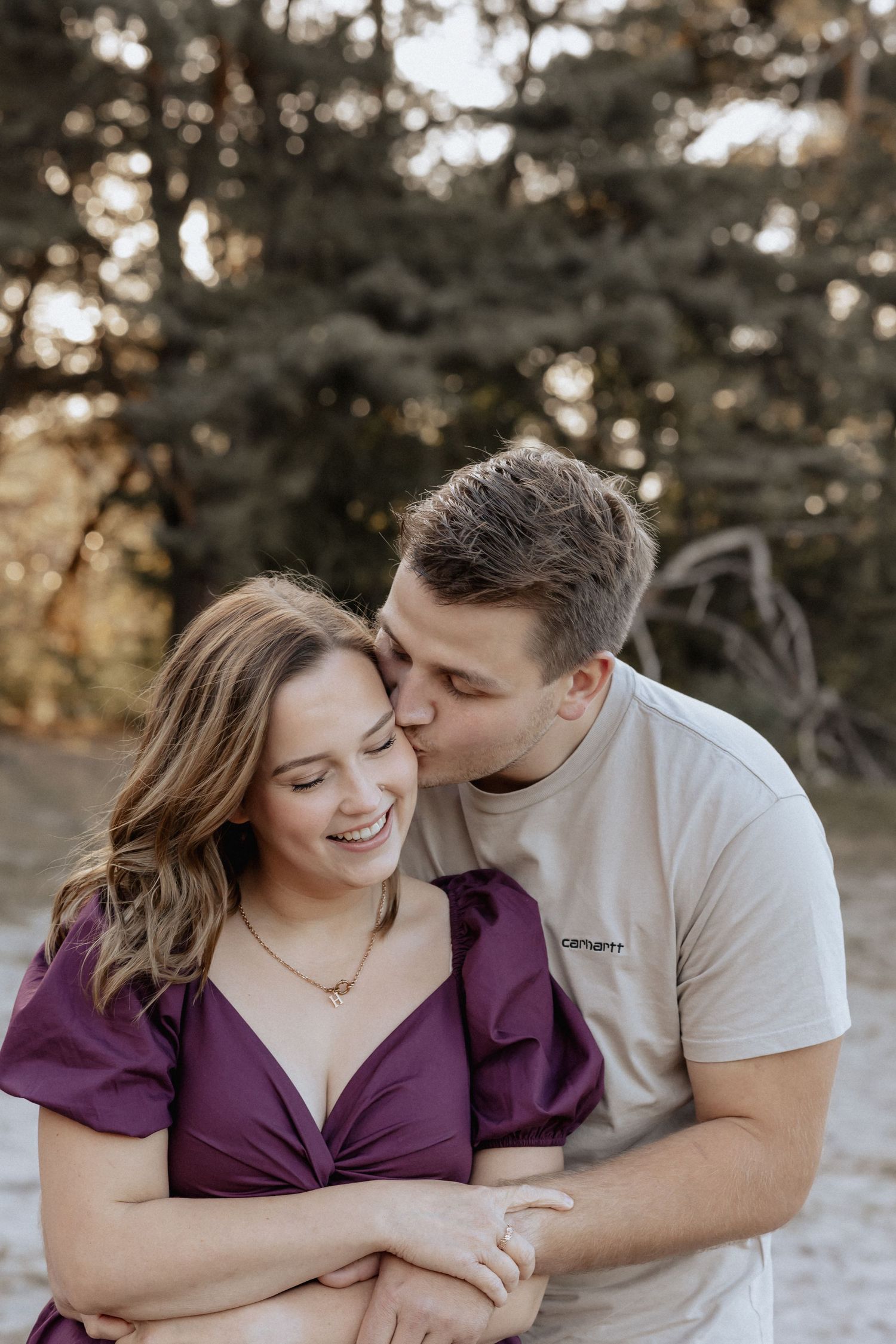 A couple shares a tender moment outdoors, man kissing woman's cheek as she smiles, wearing a purple dress.