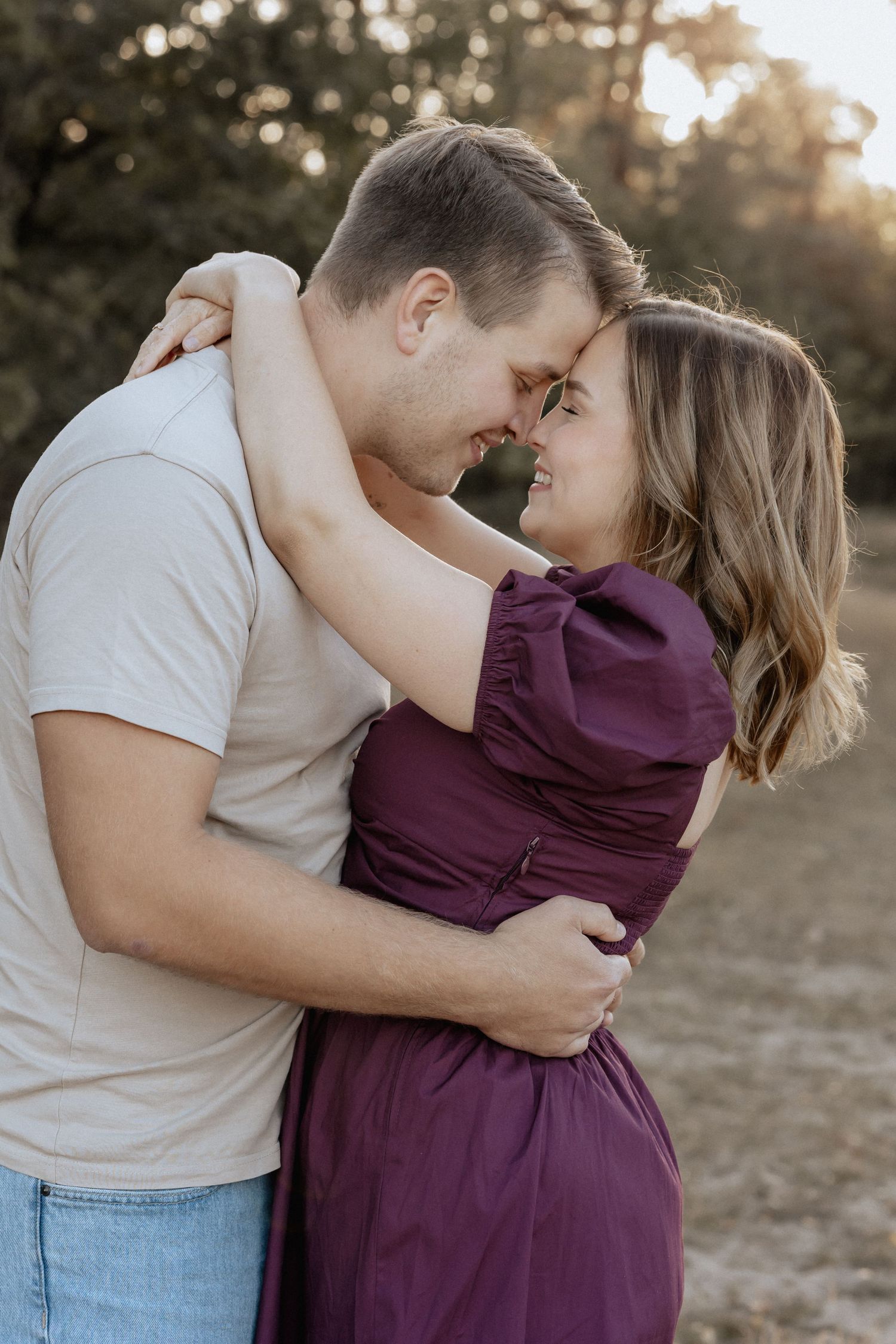A couple embraces and smiles at each other outdoors, woman in purple dress, man in white shirt and jeans.