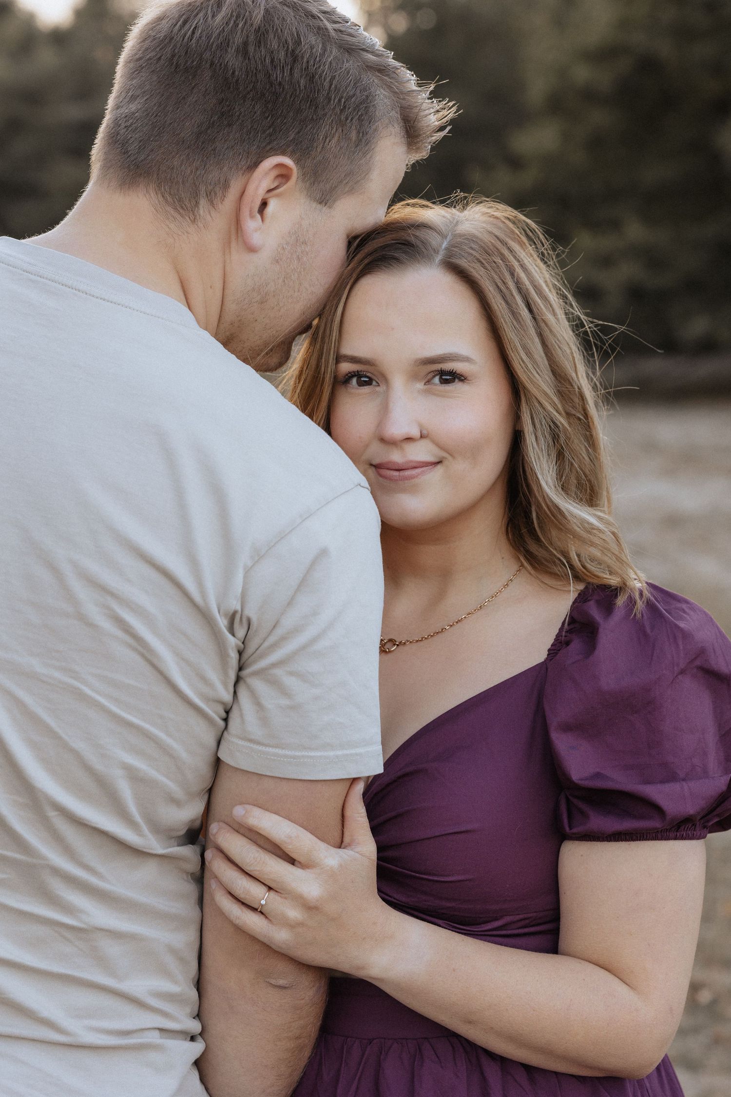A woman in a purple dress smiles at the camera while a man in a gray t-shirt kisses her forehead outdoors.