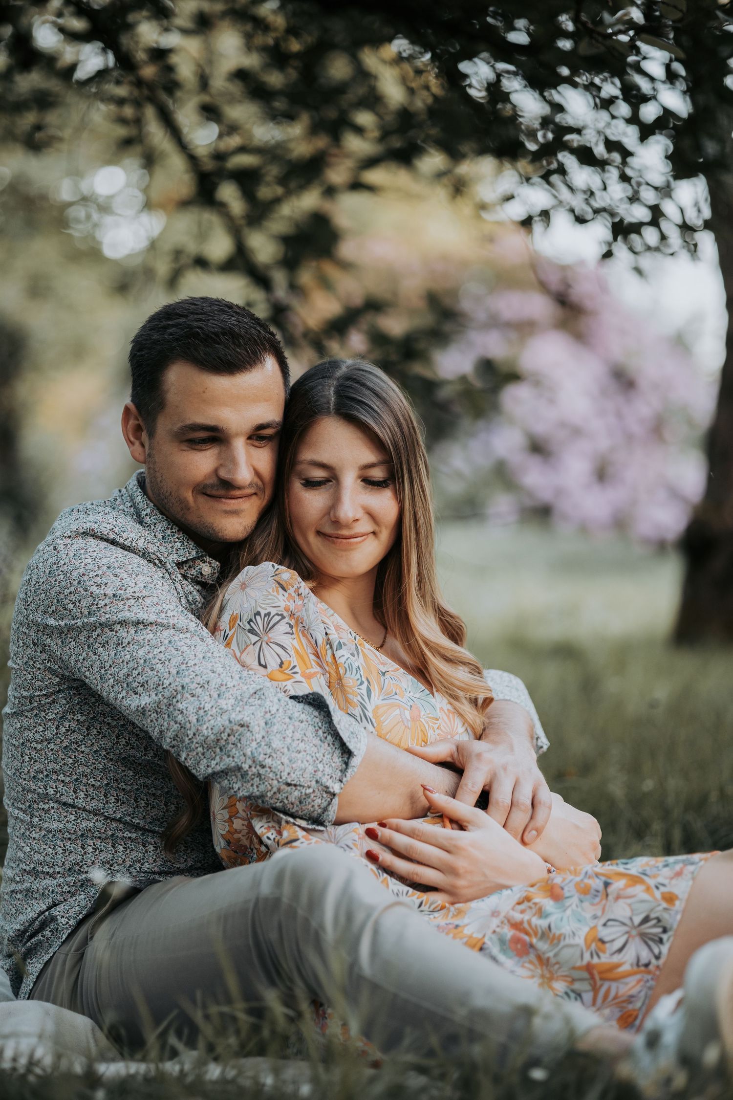 A romantic outdoor portrait of a couple cuddling on grass with cherry blossoms in the background during springtime.