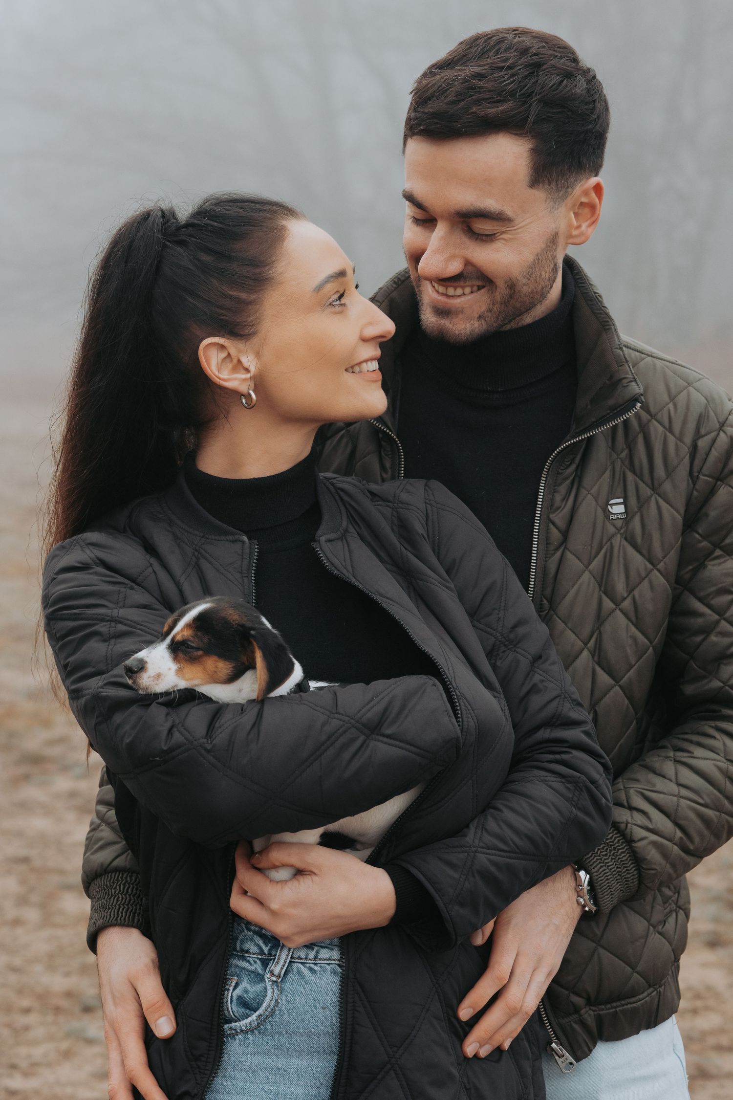 Smiling couple in quilted jackets holding a small Jack Russell terrier dog outdoors on a foggy day.
