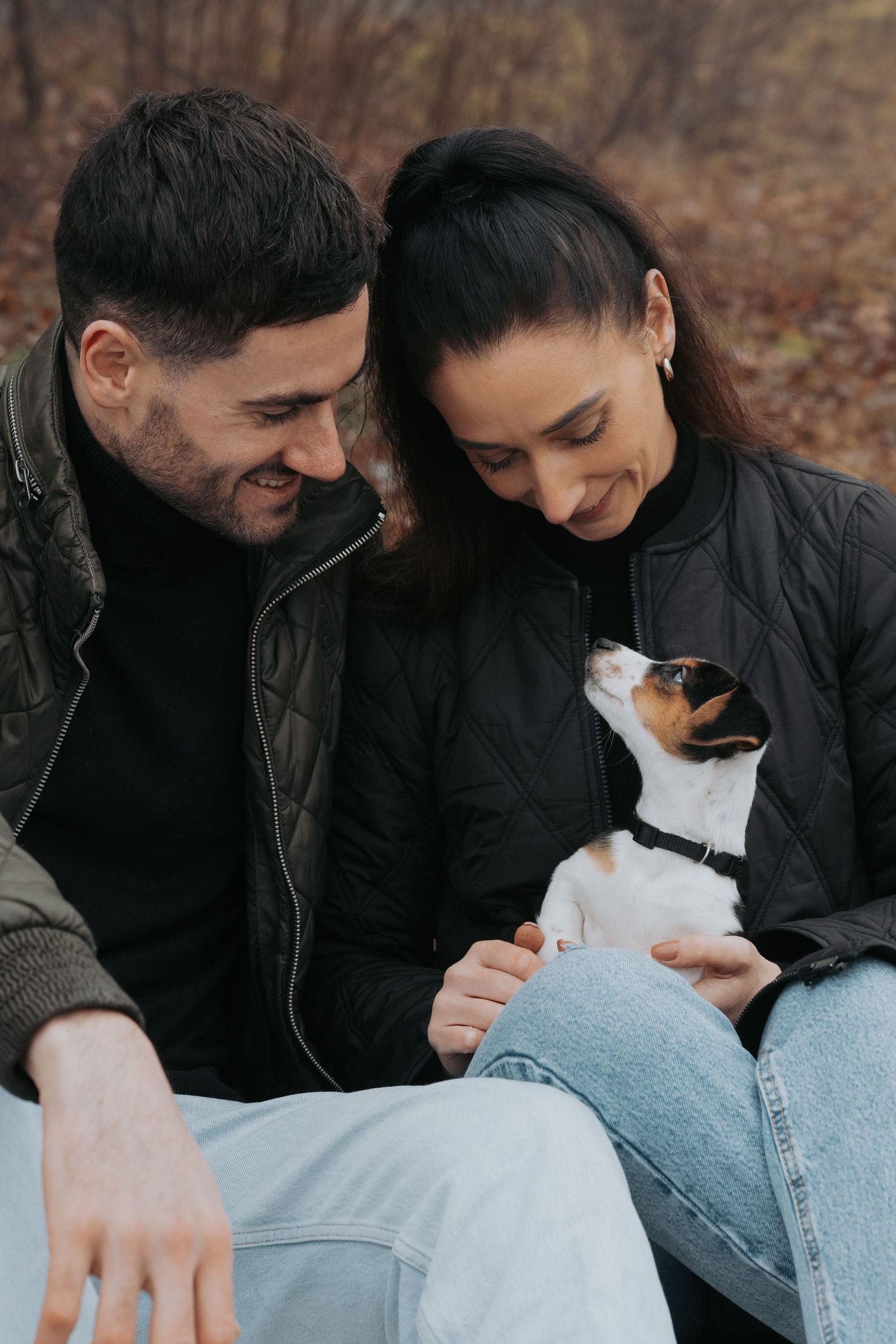 Happy couple in black jackets smiling at their small Jack Russell Terrier puppy outdoors in autumn park.