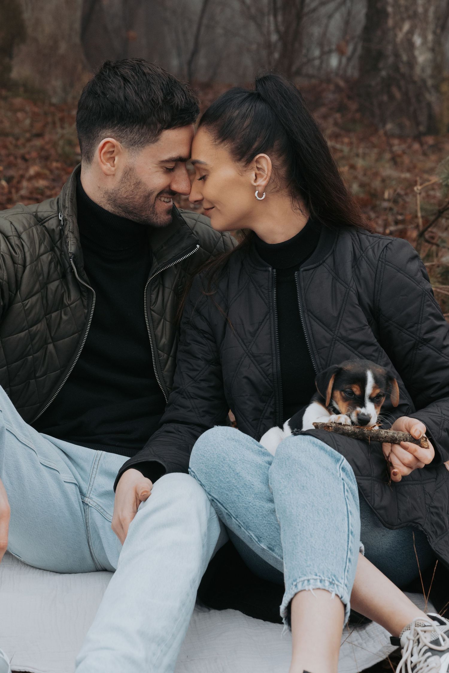 Romantic couple sitting outdoors in autumn forest, smiling at each other while holding a small beagle puppy.