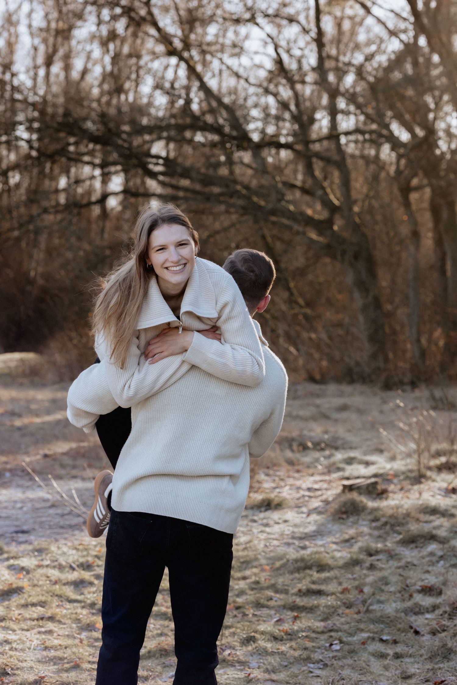 A smiling woman in a cream sweater being lifted by a man outdoors among bare trees in a romantic autumn setting.