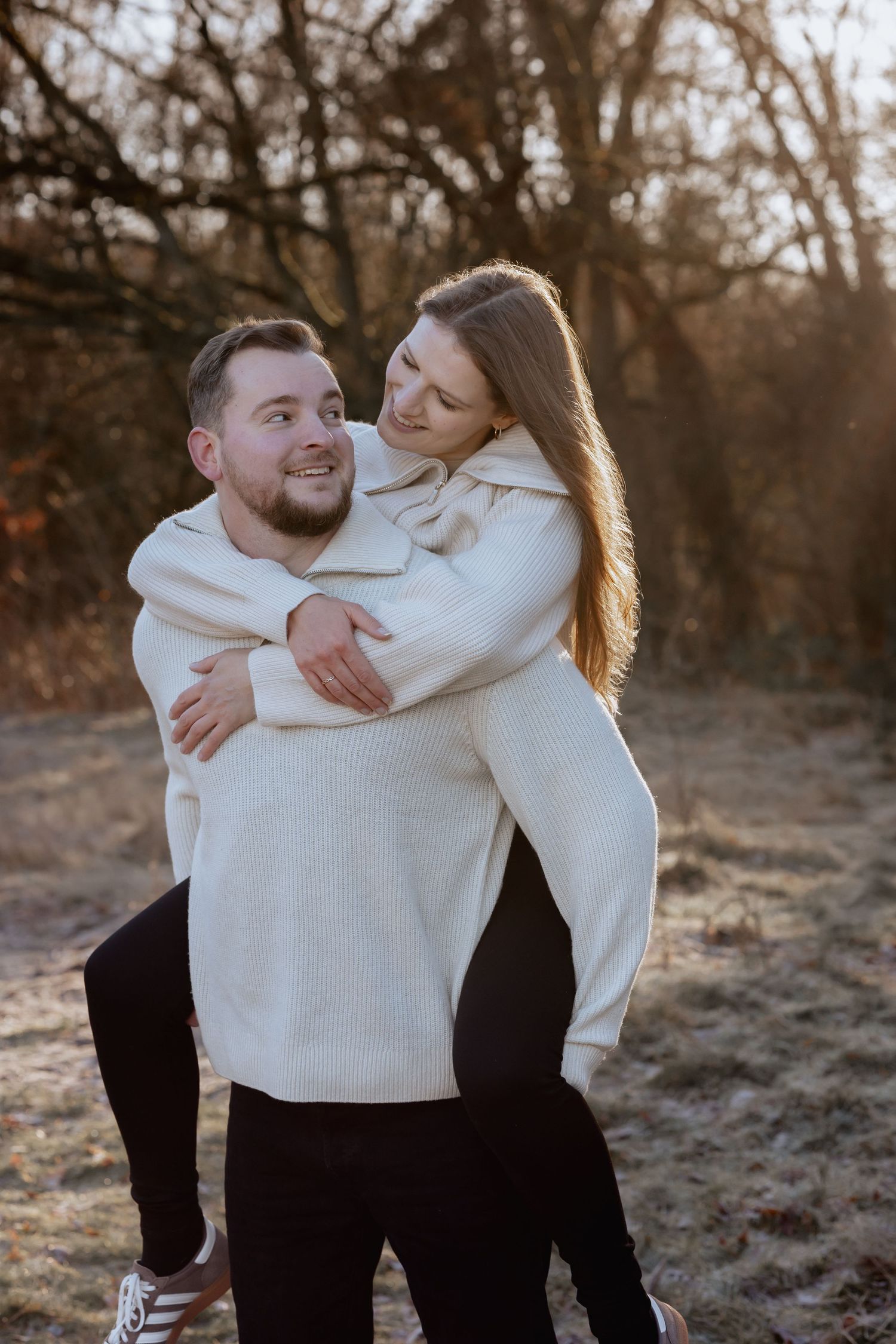 Couple in matching cream sweaters sharing a piggyback ride outdoors in a sunlit autumn park setting.