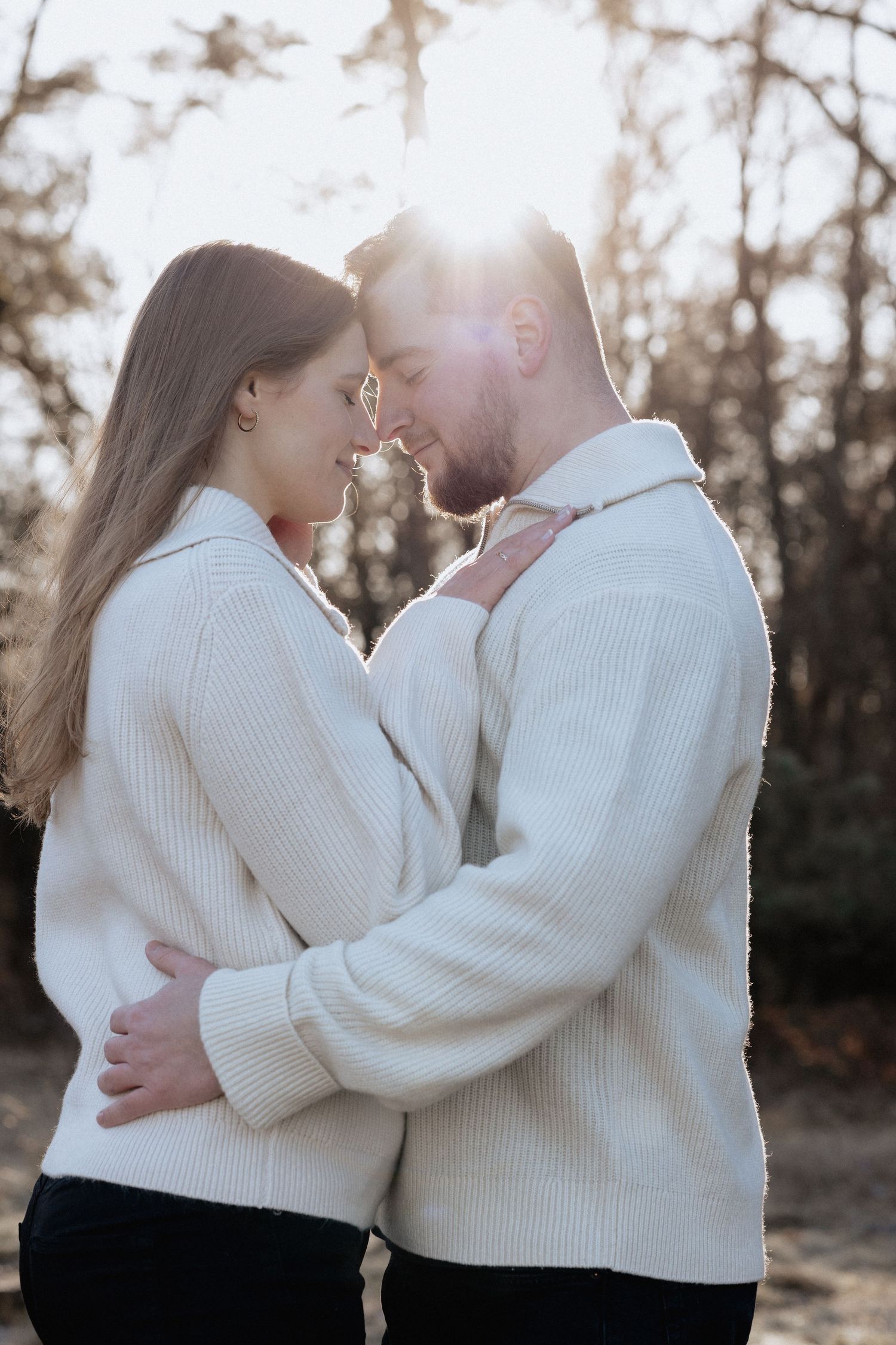 Romantic couple in matching white sweaters embracing outdoors with warm sunlight glowing behind them in a forest setting.