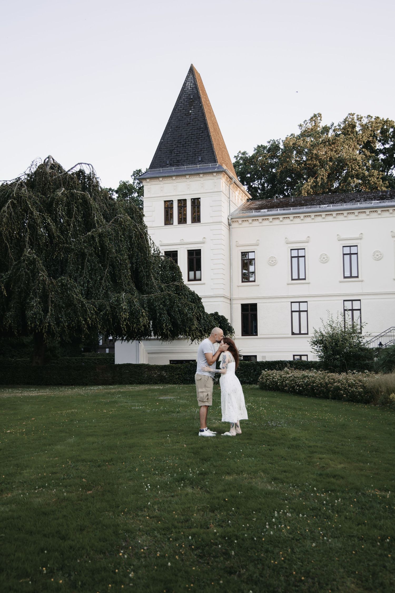 Couple embracing on green lawn in front of elegant white castle with pointed tower and weeping willow tree.