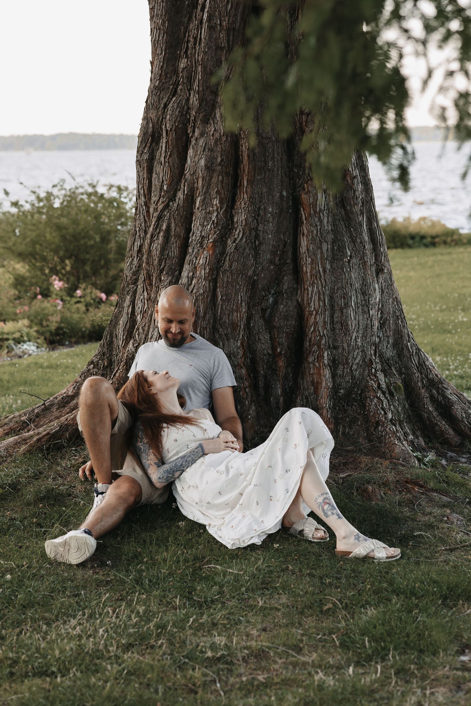 Couple sitting together against large tree trunk by waterfront, woman in white dress leaning on man in gray shirt.