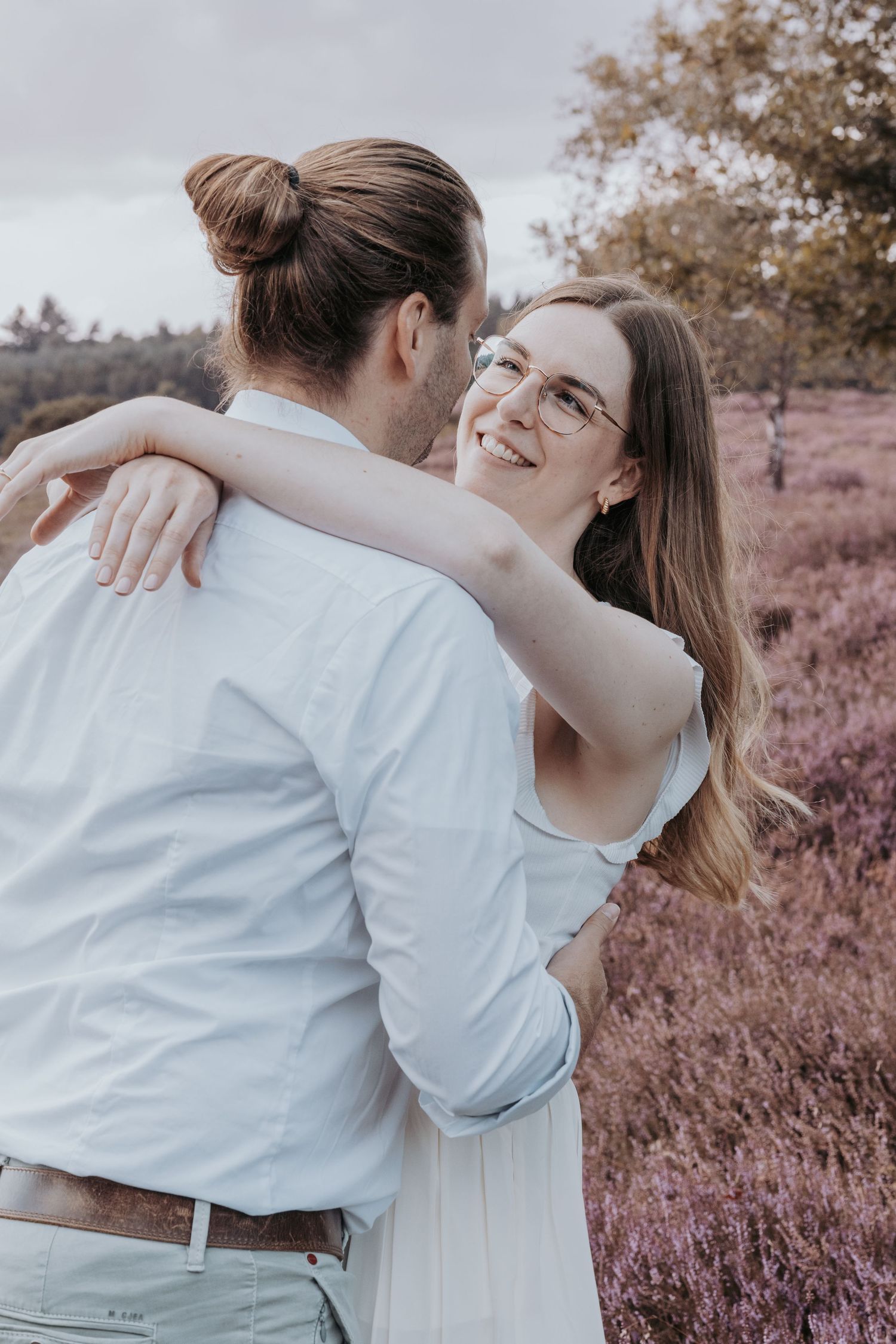 Couple embracing in a blooming heather field, smiling woman with glasses in white dress hugging man with hair bun.