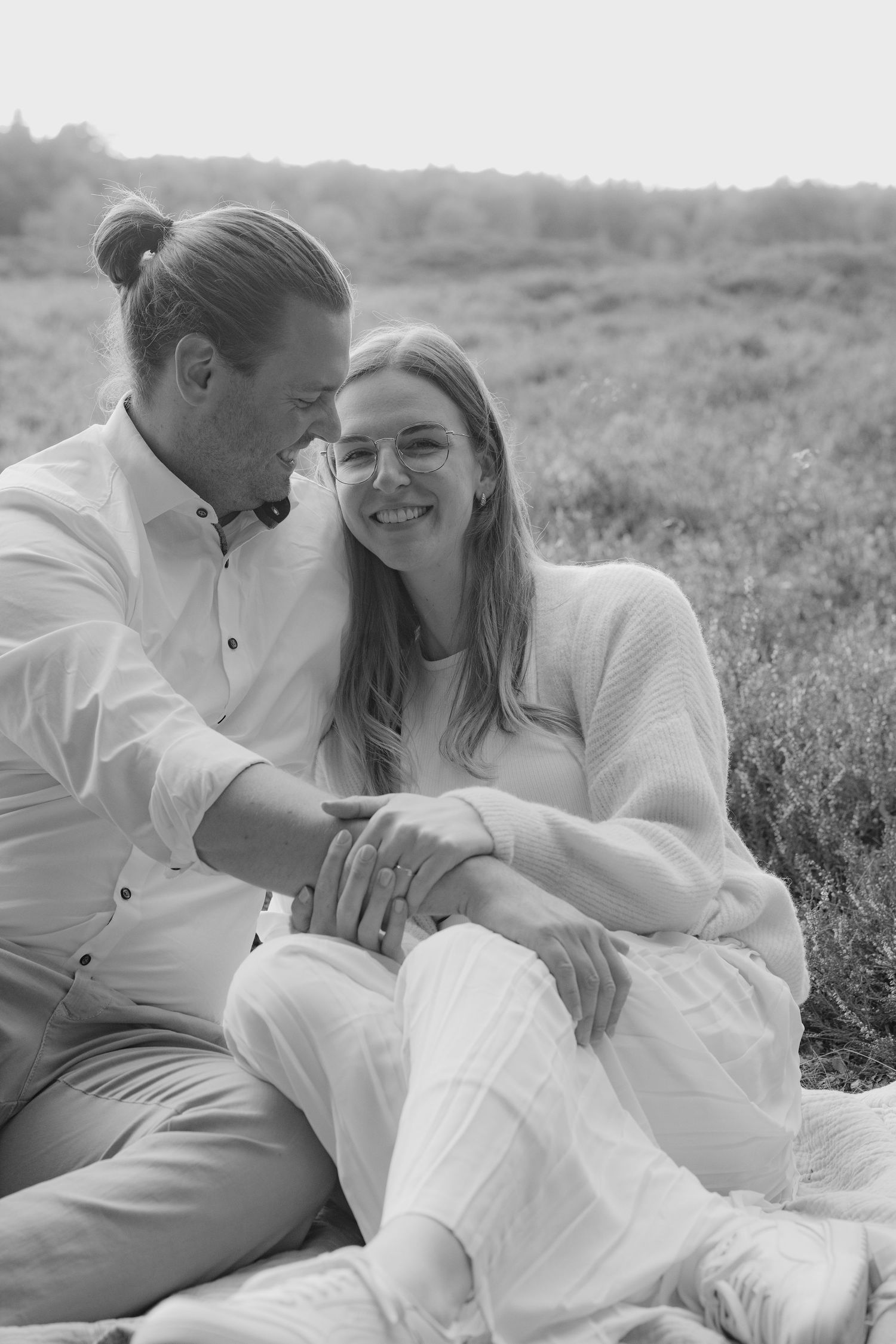 Black and white photo of a happy couple sitting together in a field, woman wearing glasses, both dressed in white.