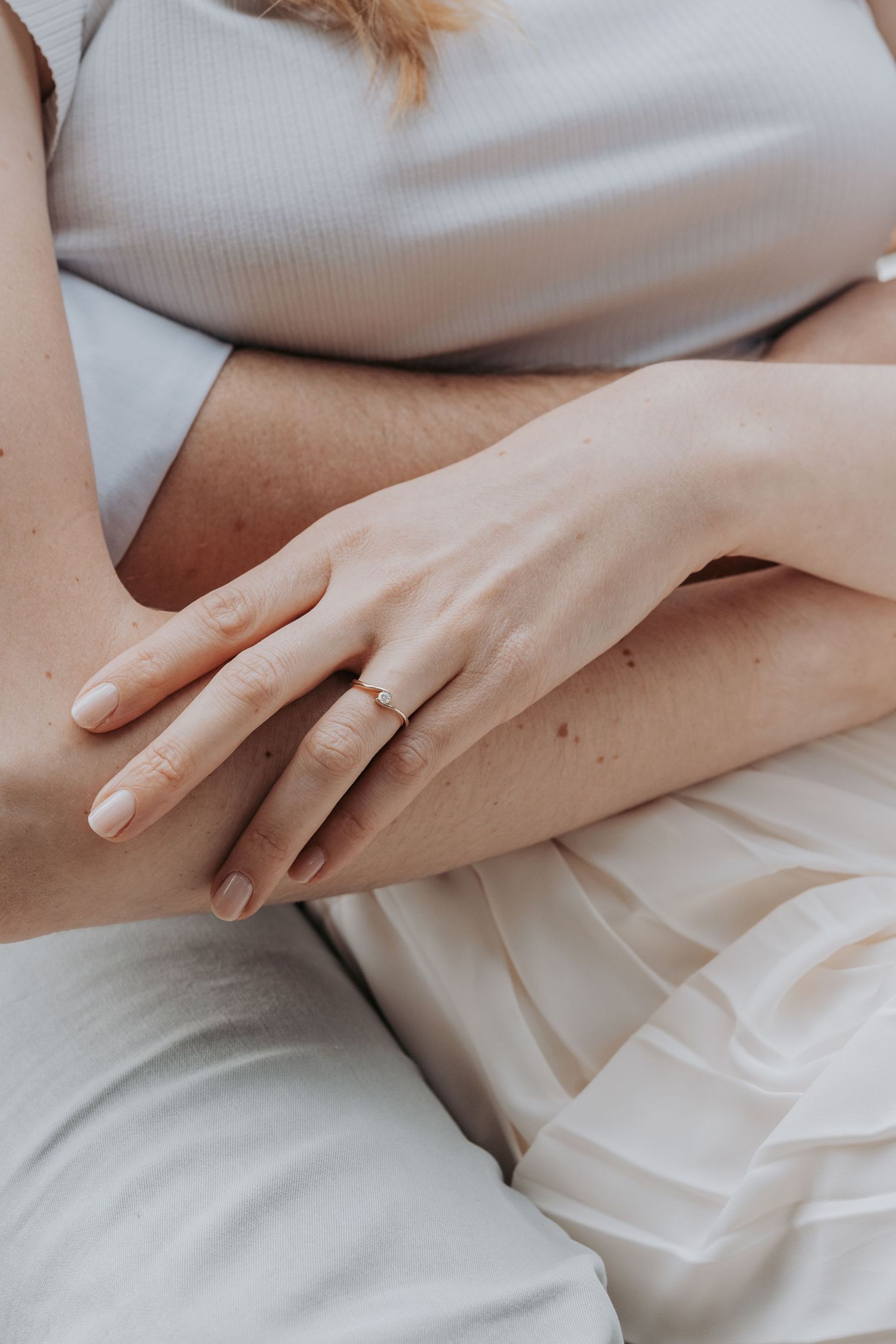 Close-up of a woman's hands resting on her lap, wearing a delicate gold ring, dressed in a soft white outfit.