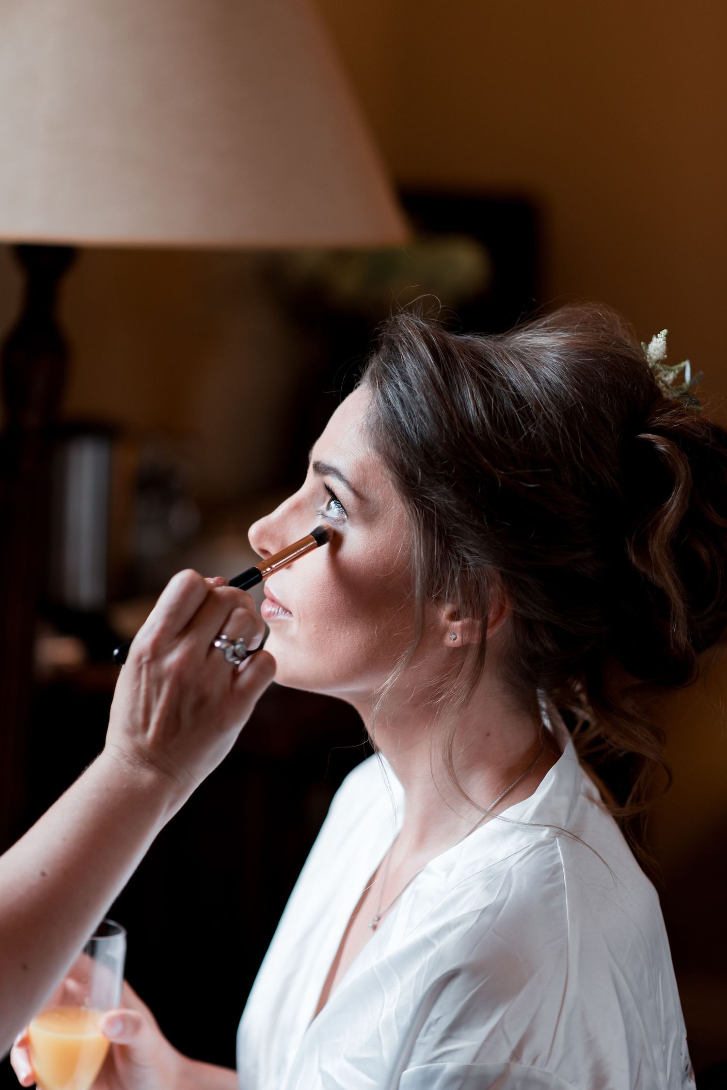 Professional makeup artist applies eyeshadow on bride during wedding preparation in white robe.