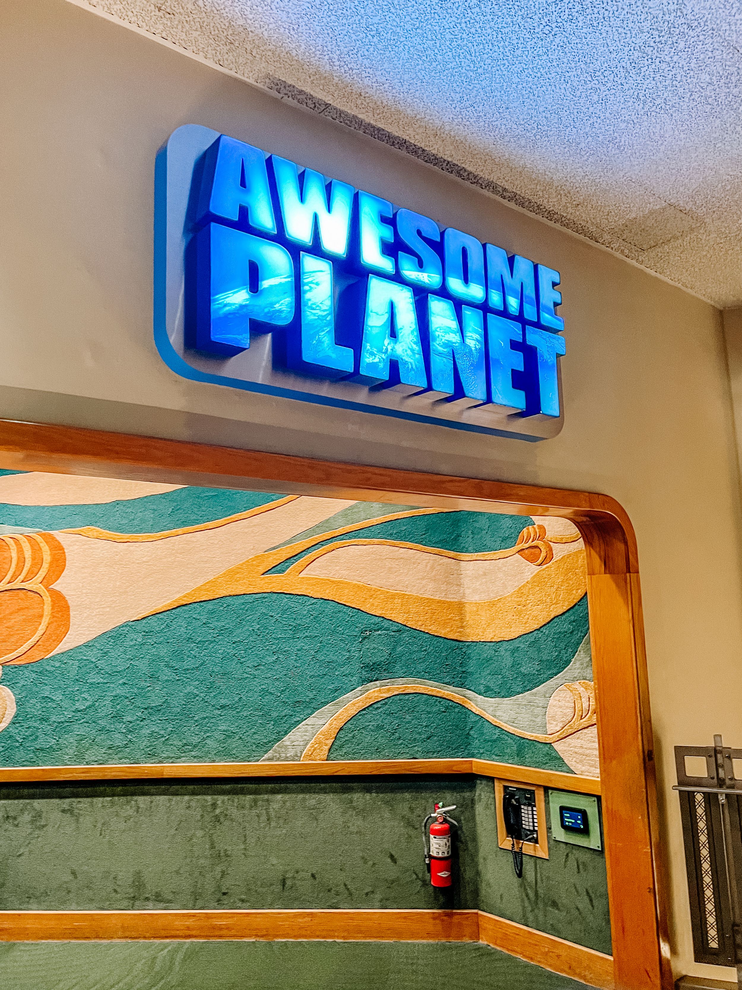 Glowing blue 'Awesome Planet' sign mounted above a retro-styled wall with wavy orange and teal artistic patterns.