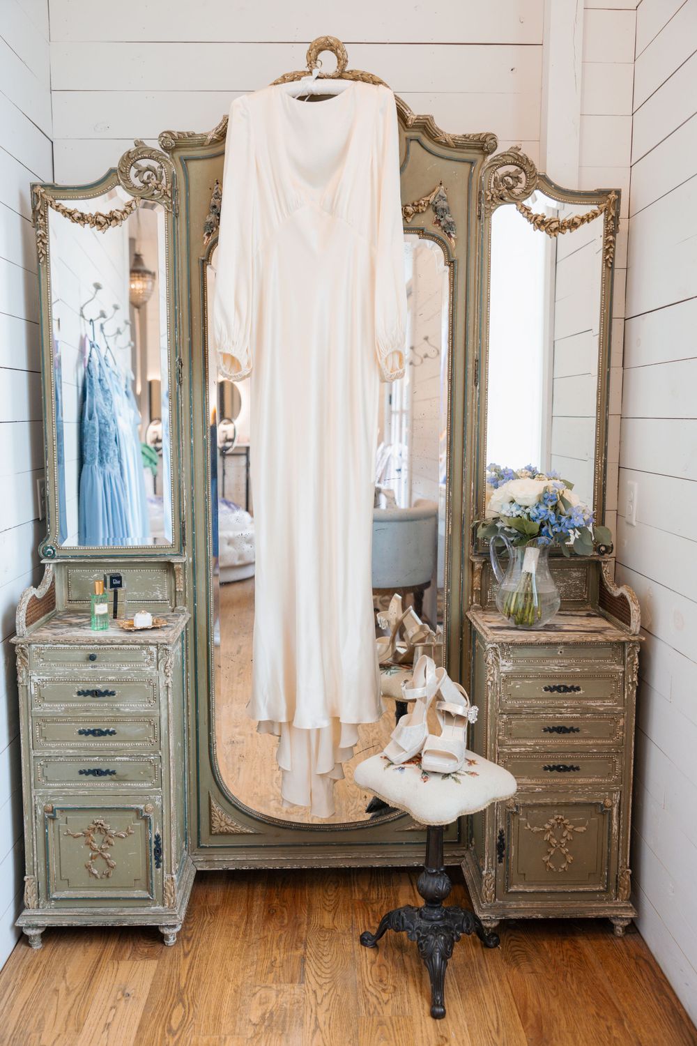 White wedding gown hanging on ornate vintage triple mirror with ballet shoes on stool in bridal suite.
