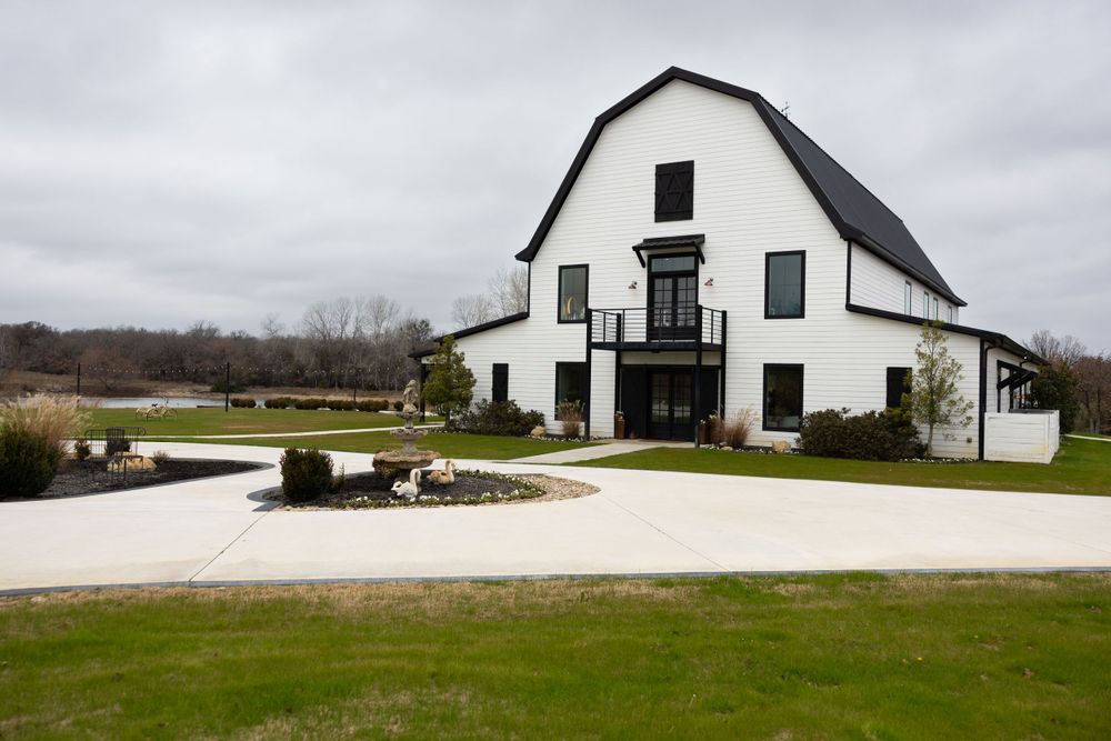 Modern white barn wedding venue exterior with black accents, balcony, circular driveway, and green lawn.