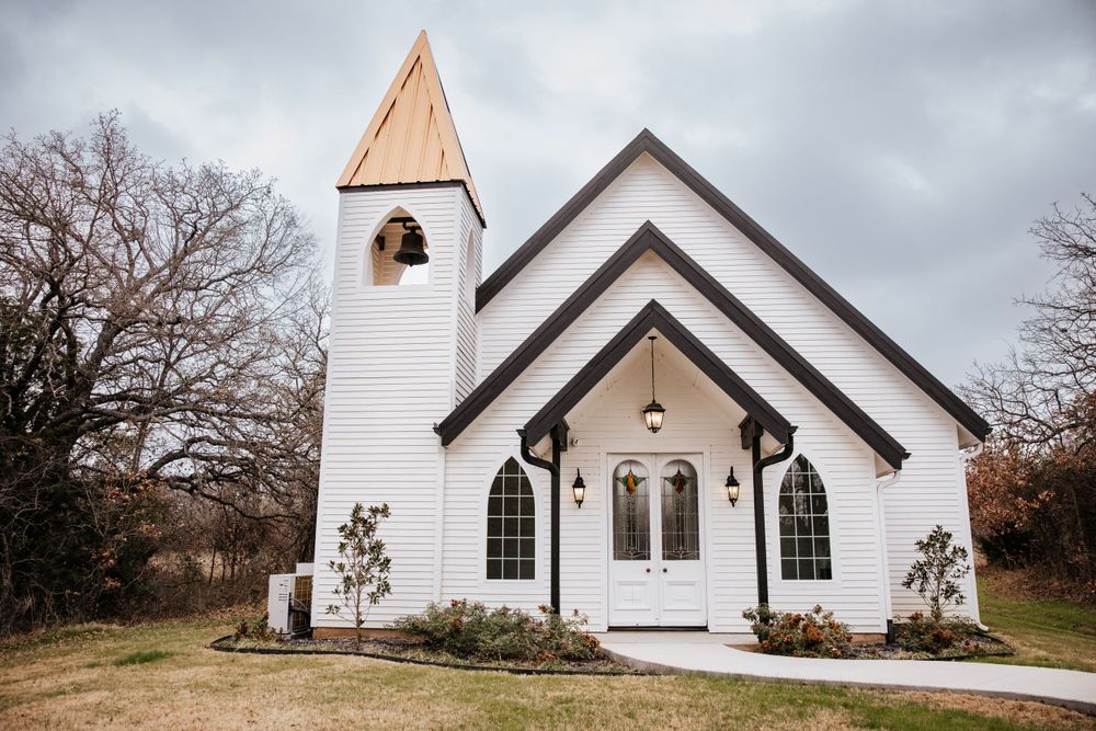 Charming white country chapel with gothic arched windows, black trim, and a wooden steeple on an overcast day.