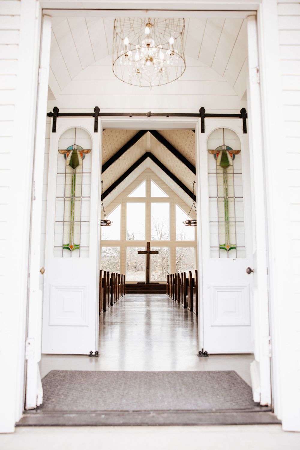 Elegant chapel interior view through open doors revealing stained glass panels, chandelier, wooden pews, and altar cross.
