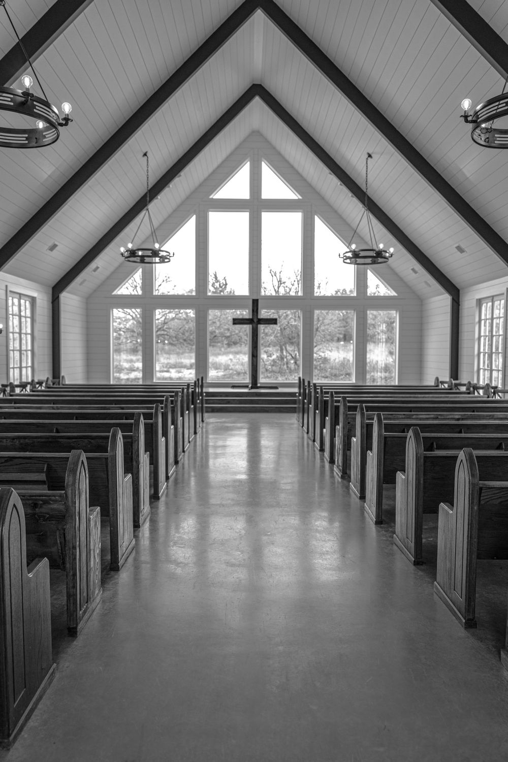 Black and white chapel interior with vaulted ceiling, wooden pews, chandeliers, and large cross window at the altar.