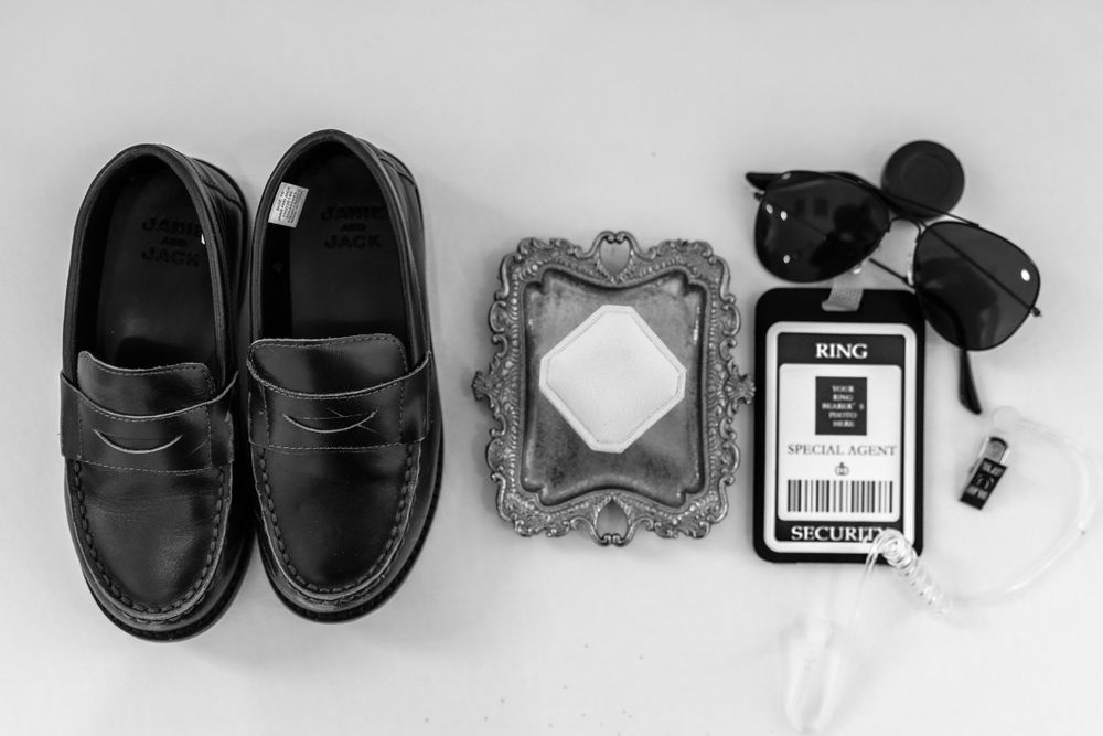 Black and white flat lay of groom wedding accessories including loafers, sunglasses, ring box, and boutonniere.