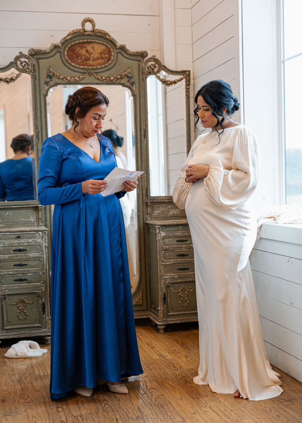 Bride in white satin gown reading a letter beside woman in royal blue dress near vintage ornate mirror.