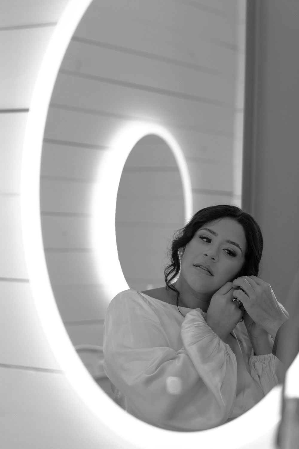 Black and white reflection of an elegant bride adjusting her earrings in a round illuminated mirror.